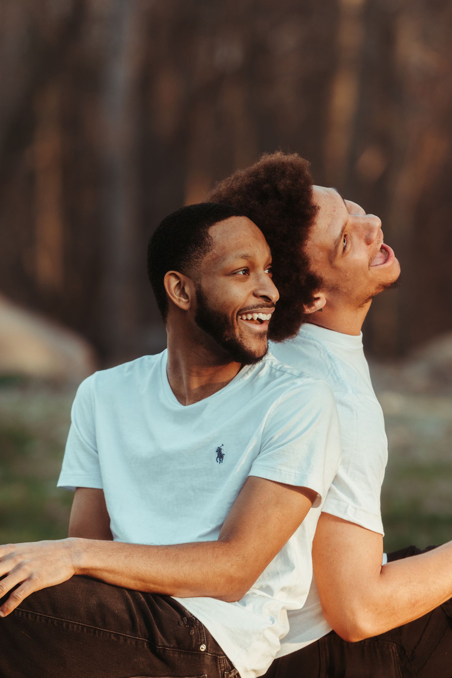 Two smiling men in light blue t-shirts sit back to back outdoors, laughing joyfully with their heads tilted up in warm golden light.