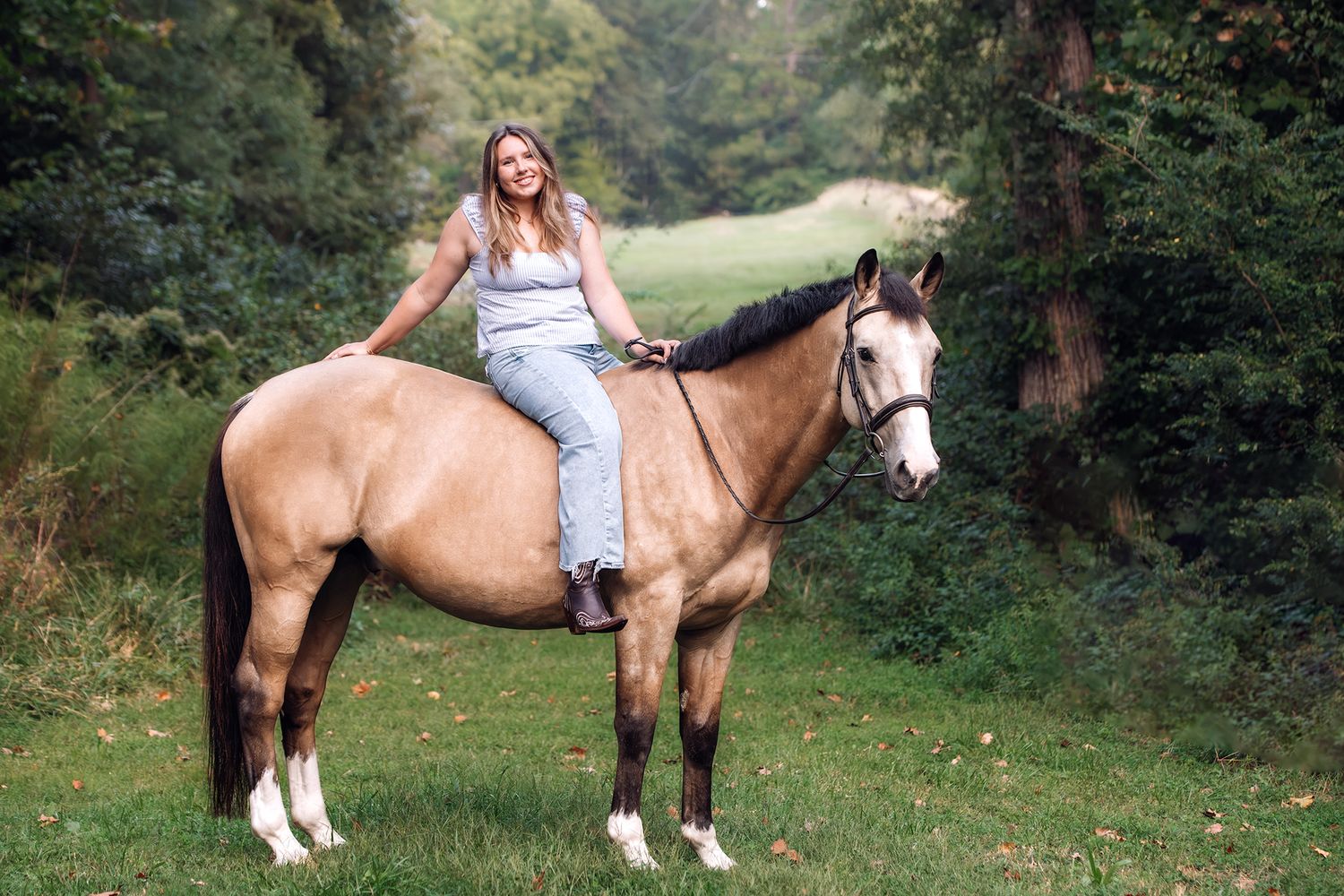 Smiling woman in gray outfit rides a buckskin horse bareback in a lush green wooded area.