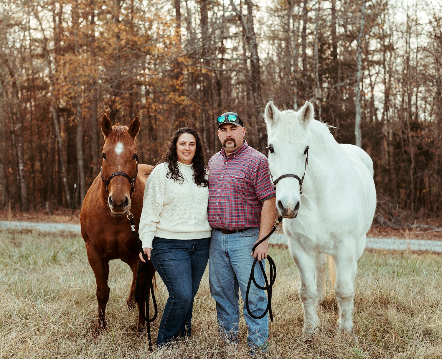 Couple standing between a chestnut and white horse in a field with autumn trees.