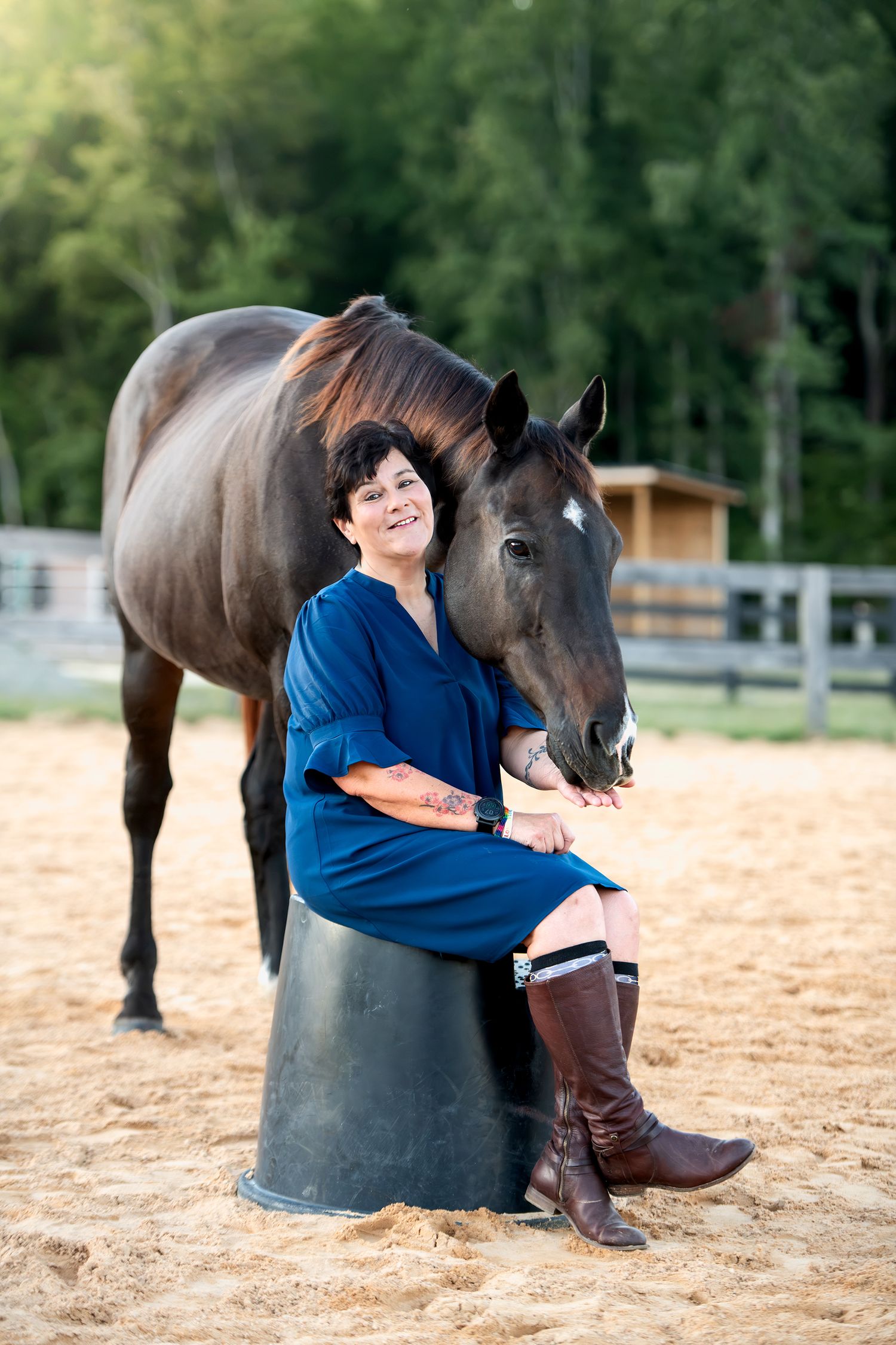Woman in blue dress sitting on bucket beside dark brown horse in sandy arena.