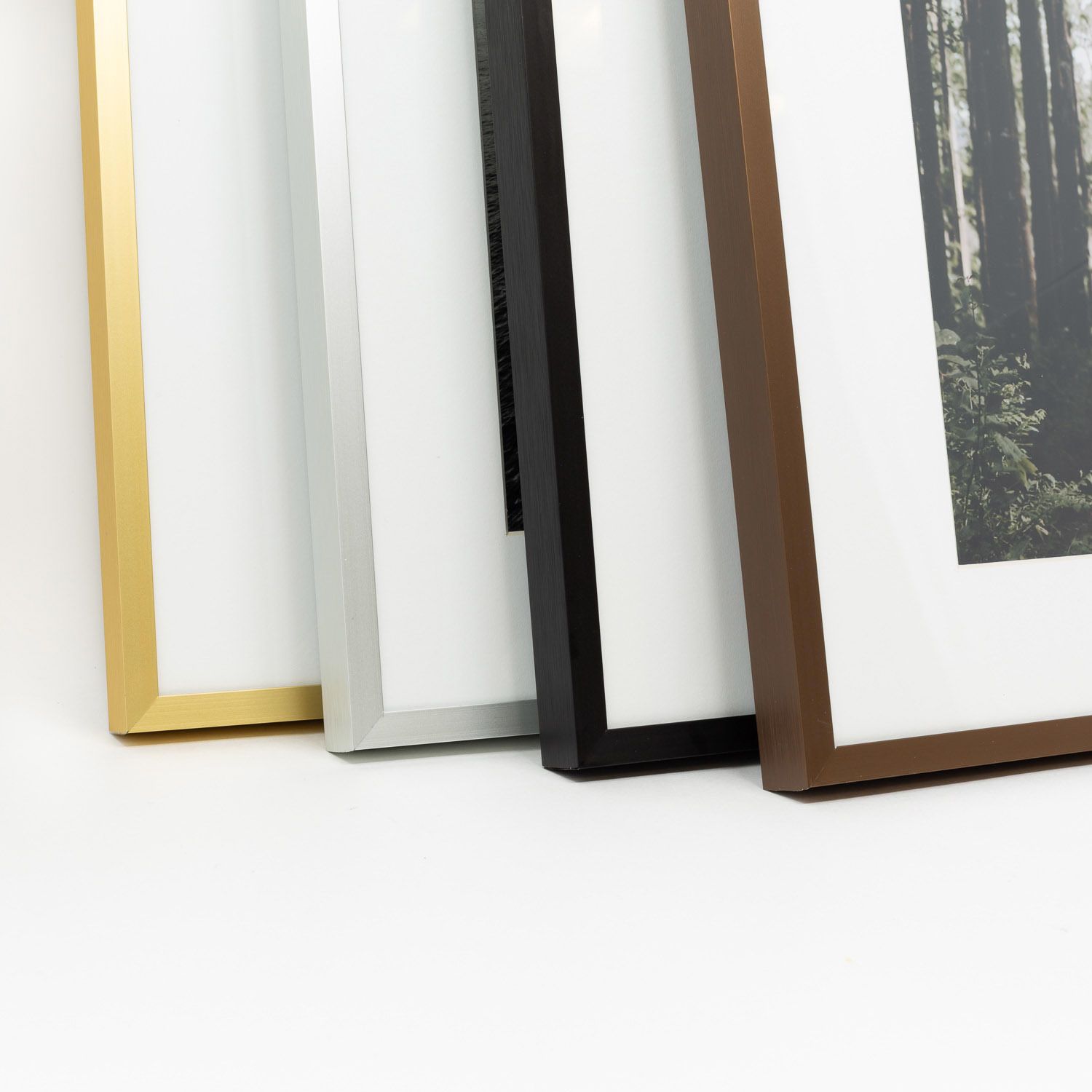Four thin modern picture frames in gold, silver, black, and walnut brown finishes leaning against a white wall with white mats.