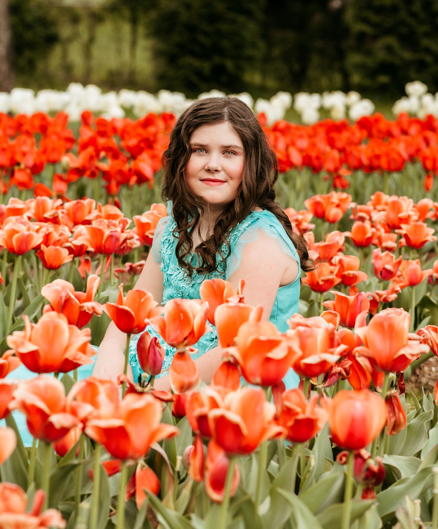 Smiling young woman with long dark wavy hair in a teal dress stands among vibrant orange and red tulips in a blooming flower field.