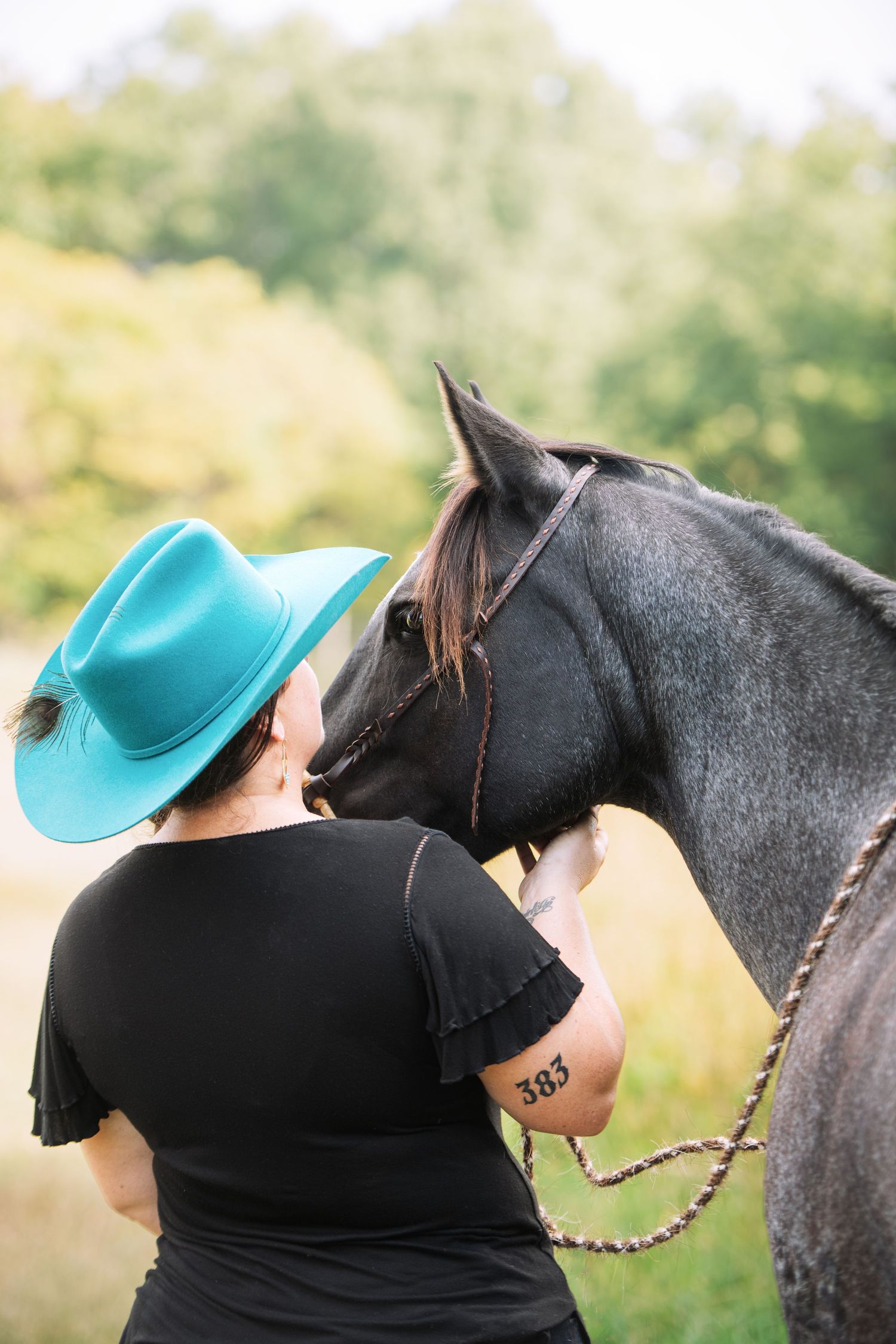 Woman in a turquoise cowboy hat and black top stands close to a gray horse in a bridle and rope halter outdoors, surrounded by green trees.
