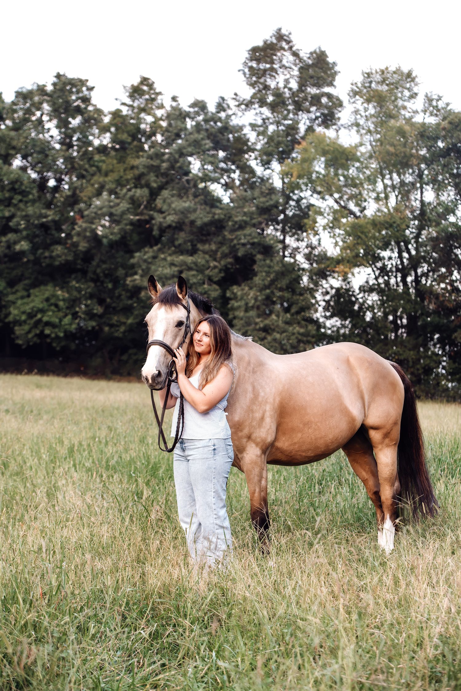 Young woman in jeans standing beside a buckskin horse in a green grassy field.