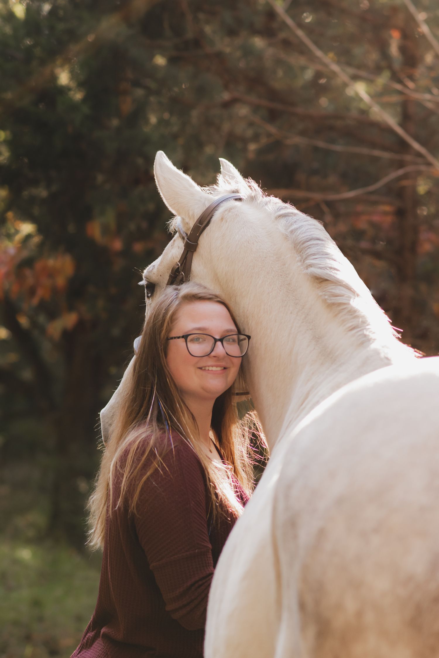 Smiling young woman with glasses posing closely with white horse outdoors in autumn.