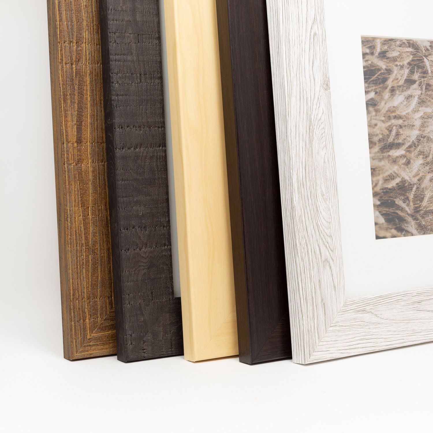 Five wood picture frames in walnut, dark espresso, natural maple, black and whitewashed gray finishes leaning together on a white surface.