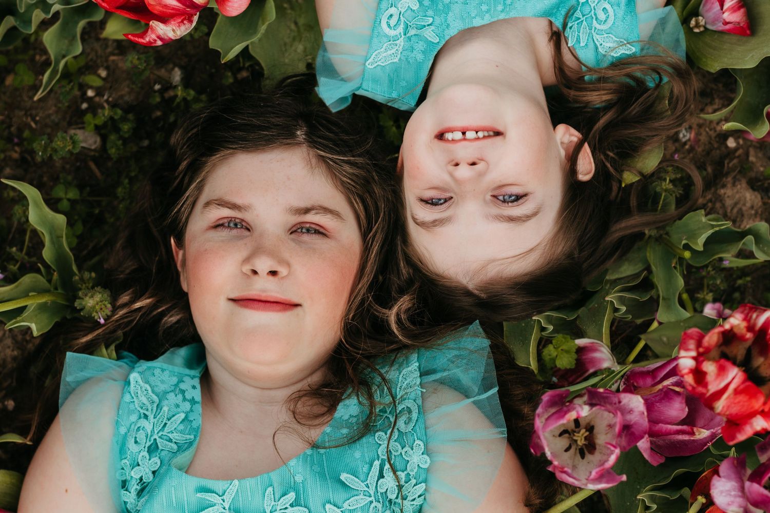 Two girls in teal dresses lying among colorful tulips smiling up at the camera.
