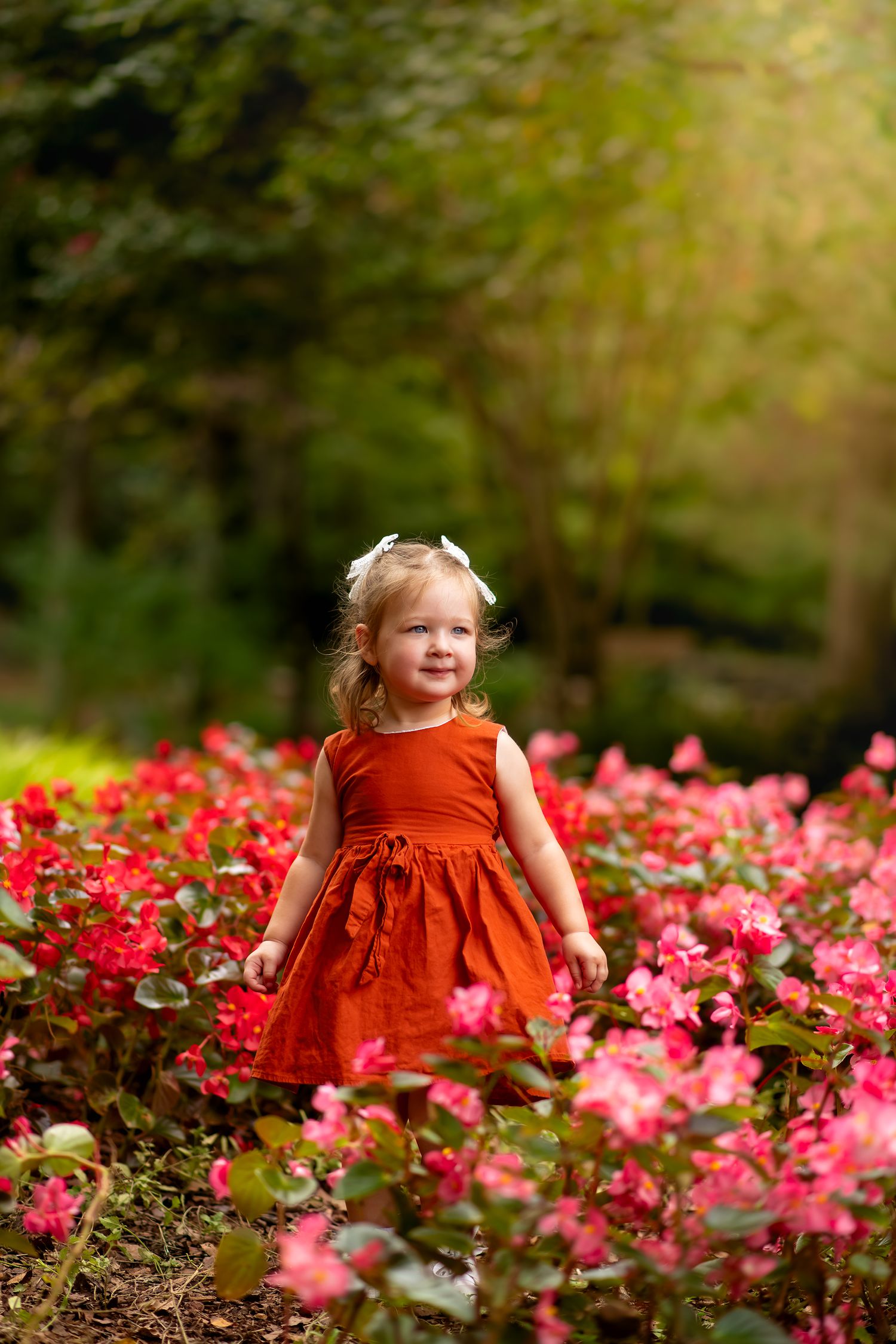 Smiling toddler girl in orange dress with white bow standing in vibrant pink flower garden.