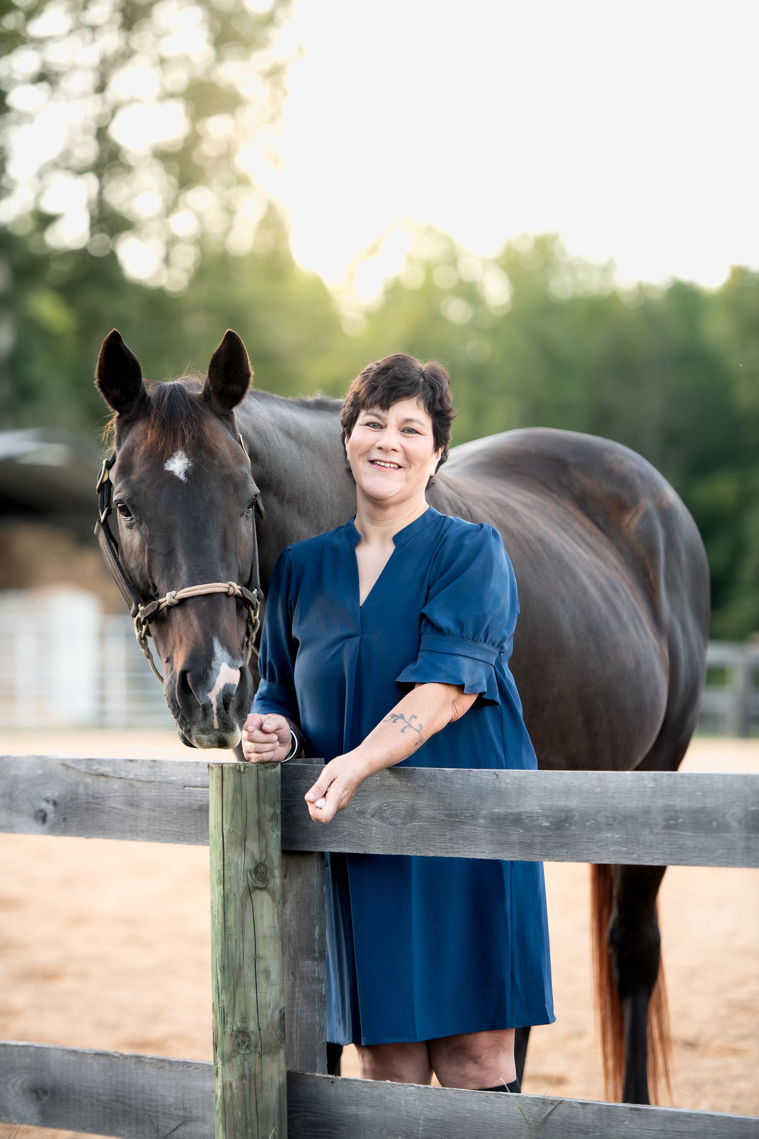 Woman in blue dress smiles beside a dark horse leaning over a wooden fence at golden hour.