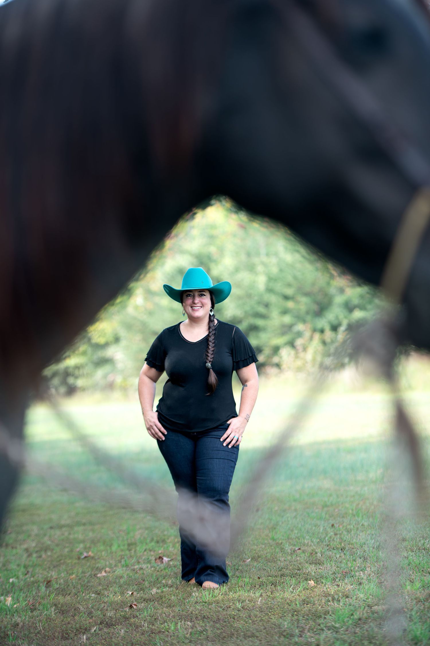 Woman in a teal cowboy hat and black shirt with a long braid smiles on a grassy field, framed by a blurred horse in the foreground.