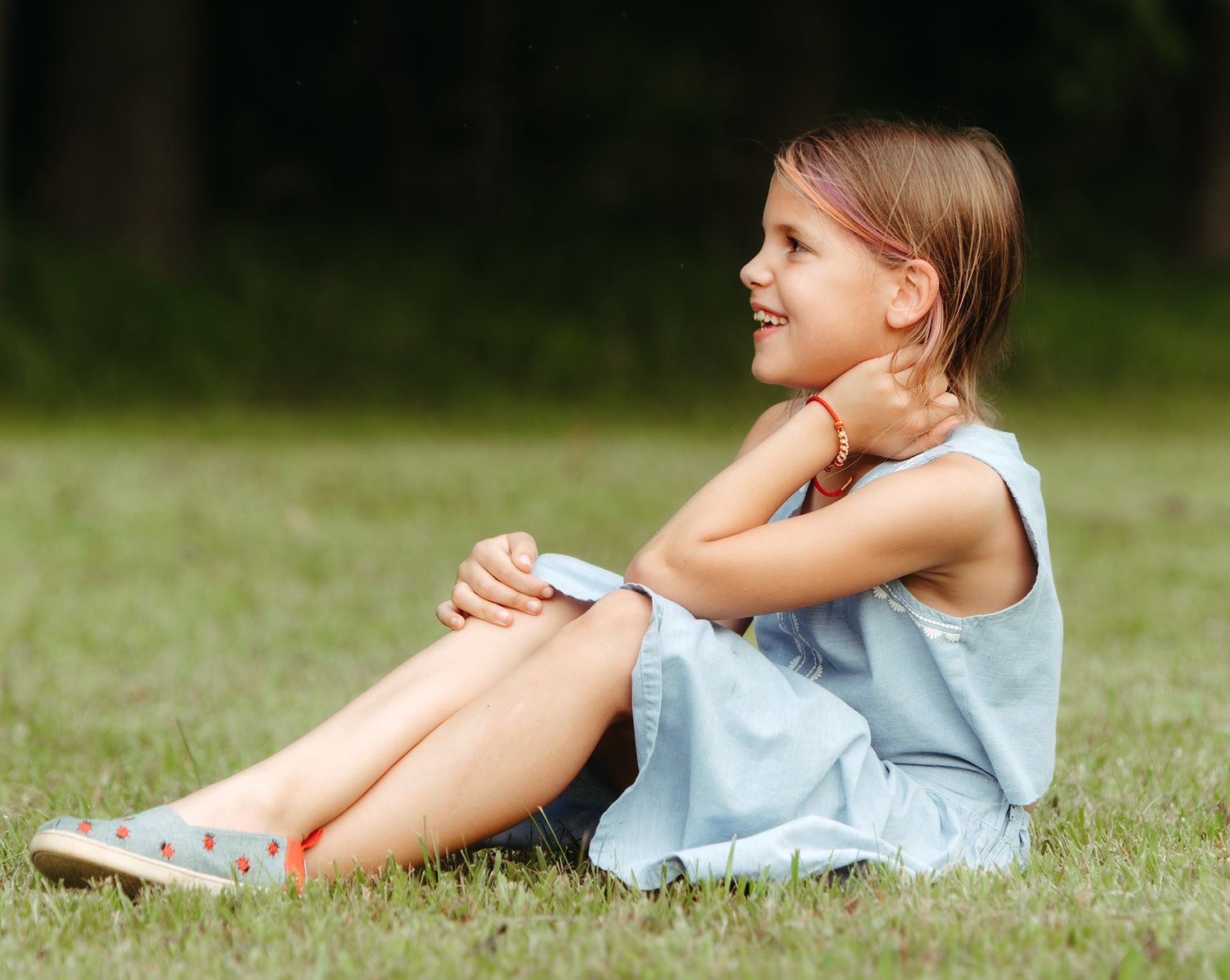 Young girl in light blue dress sitting on grass in park, smiling sideways.