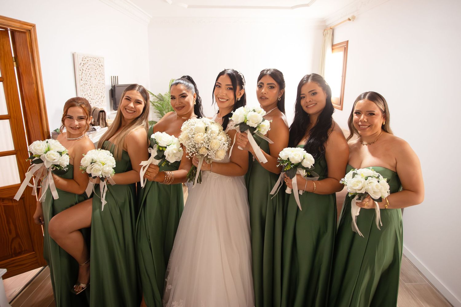 Bridesmaids in olive green dresses pose with white bouquets in a bright indoor setting.