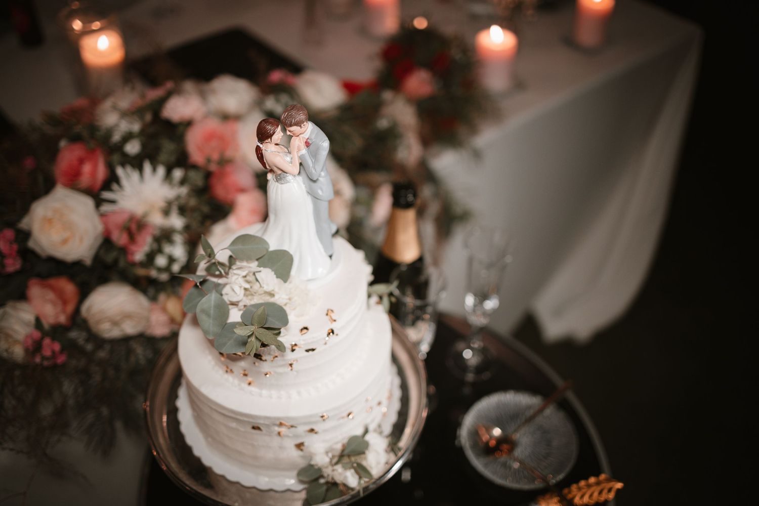 White wedding cake topped with a miniature couple figurine surrounded by pink flowers and candles.