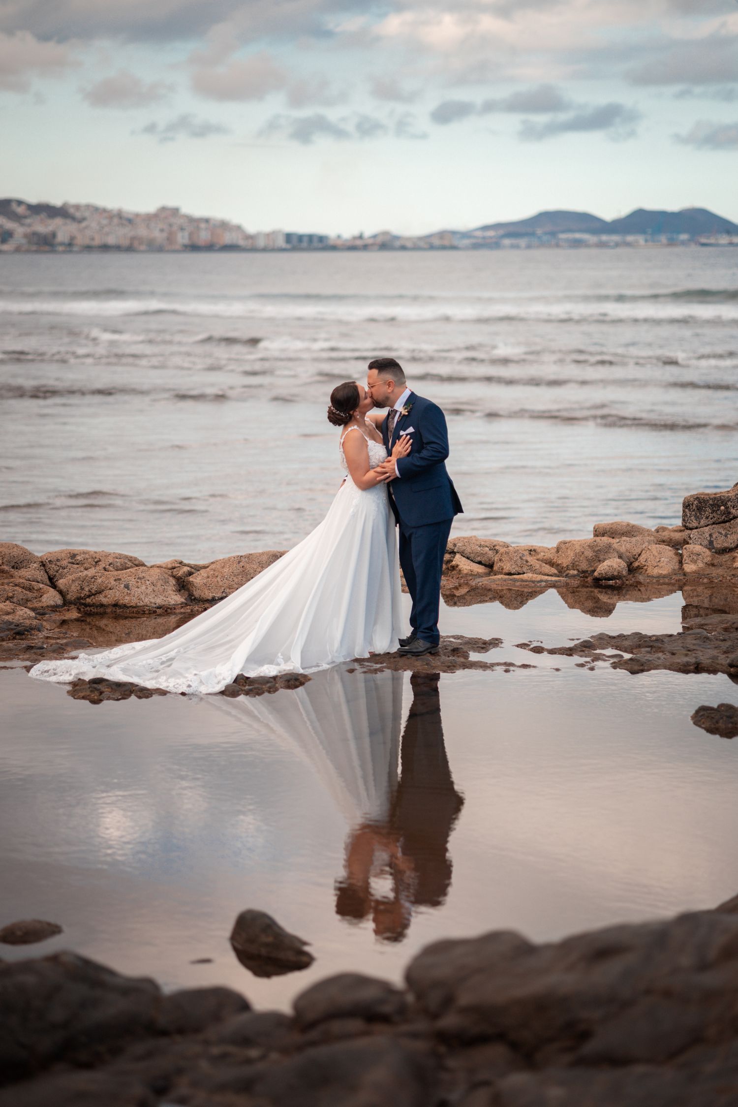 A couple in wedding attire stands on a rocky beach at sunset with waves crashing behind them.