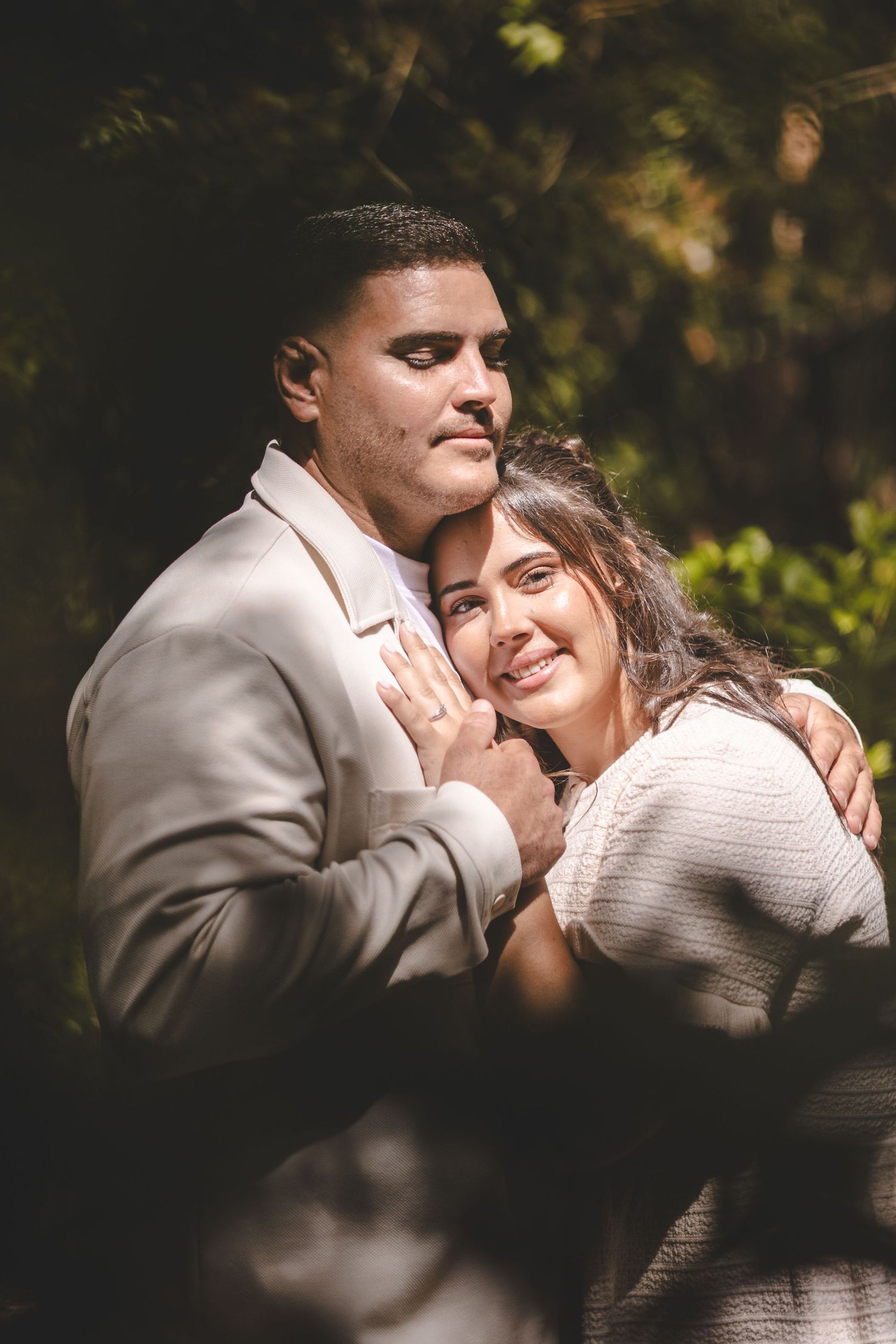 Couple embracing outdoors, man in gray suit hugging smiling woman in white sweater, surrounded by lush green foliage.