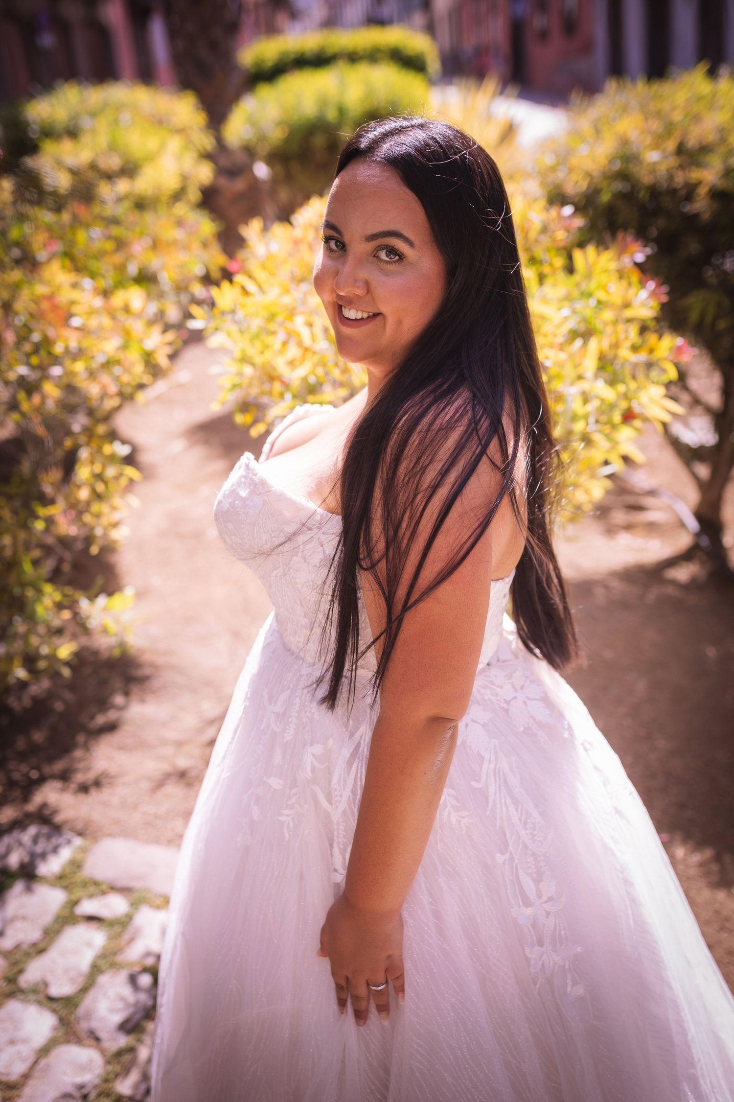 Bride in flowing white gown poses in sunlit garden path surrounded by lush foliage.