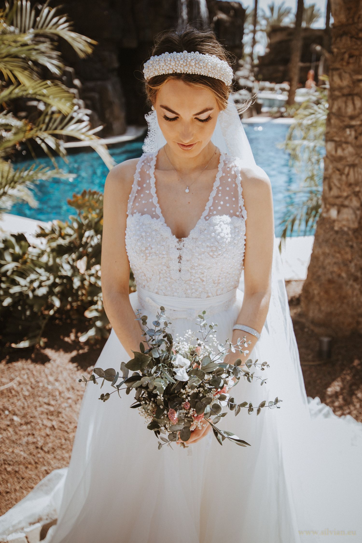 A bride in a detailed white lace gown holds her bouquet in a tropical poolside setting.
