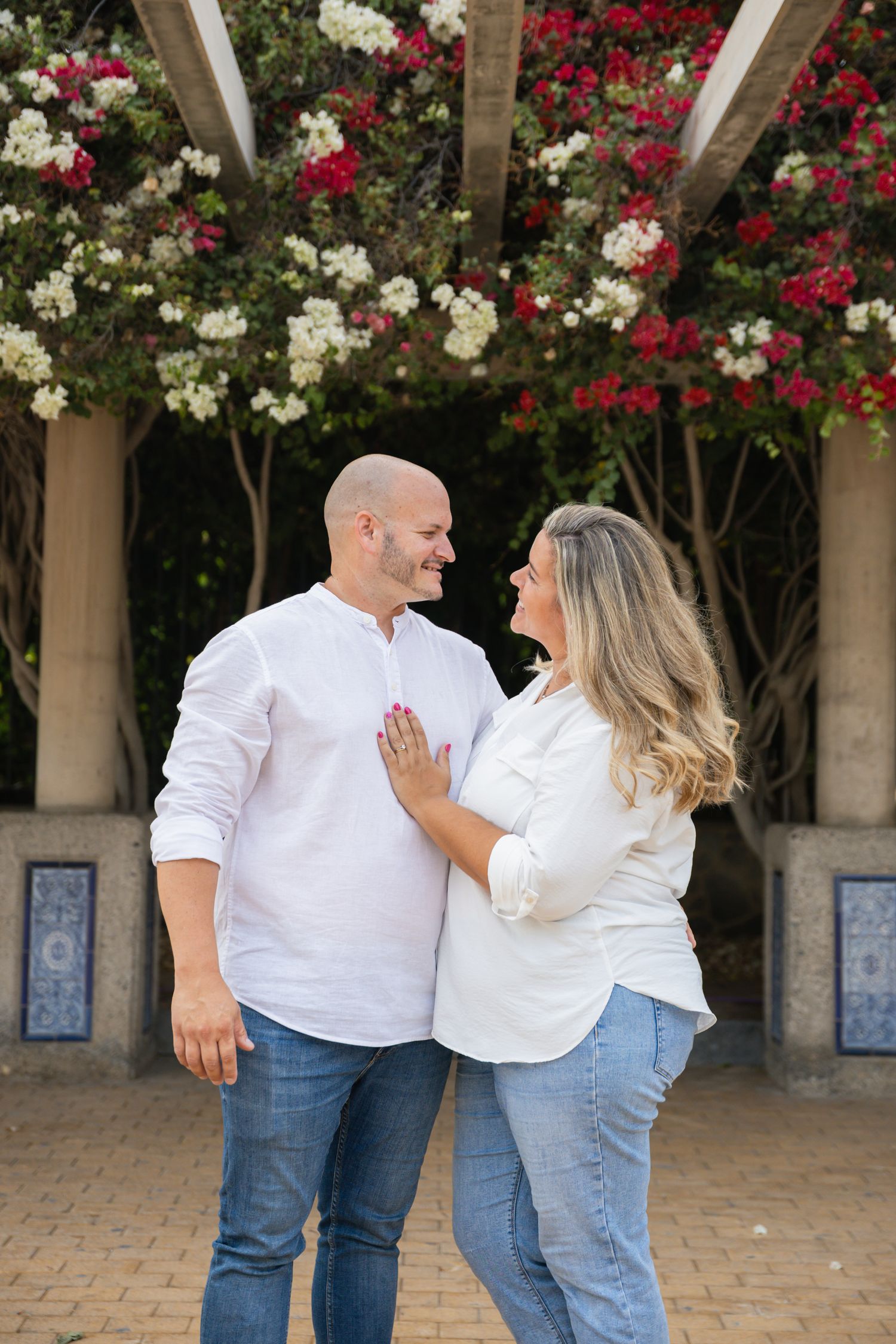 Couple in matching white tops and jeans pose playfully under flowering archway.