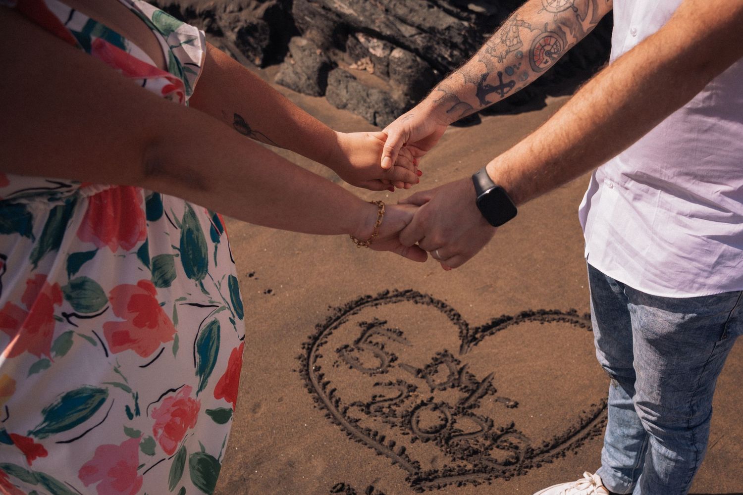 Two people drawing hearts in the sand on a beach near rocky cliffs.