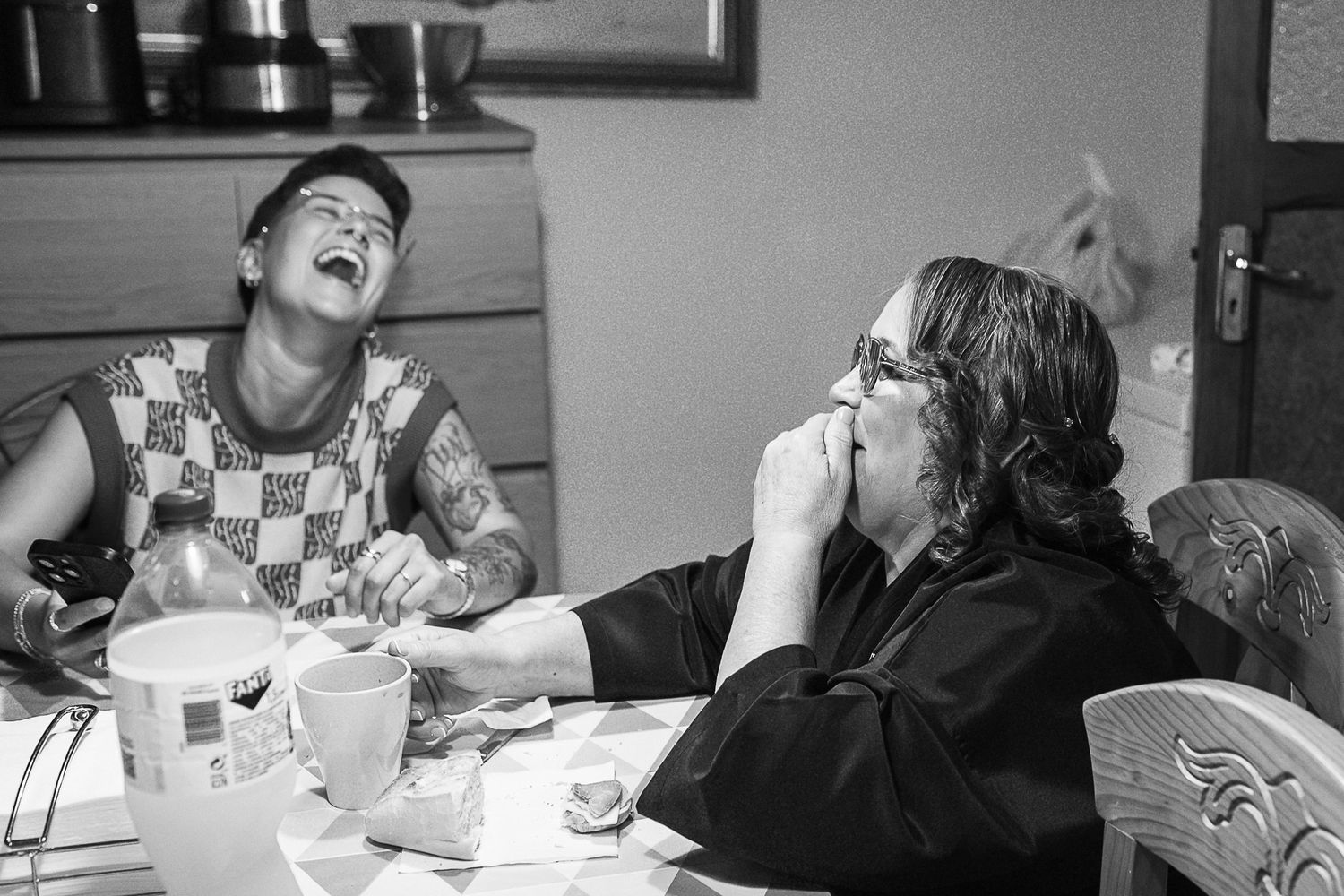 Two women laughing joyfully together at a kitchen table in a candid black and white photo.