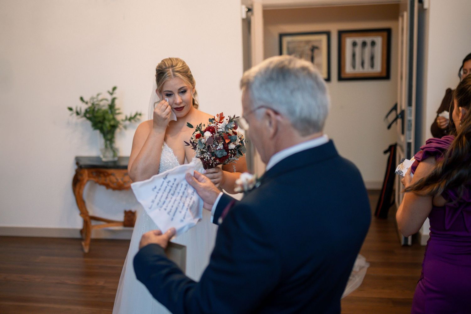 Emotional bride in white gown reads vows while holding bouquet, wiping tears during intimate wedding ceremony indoors.