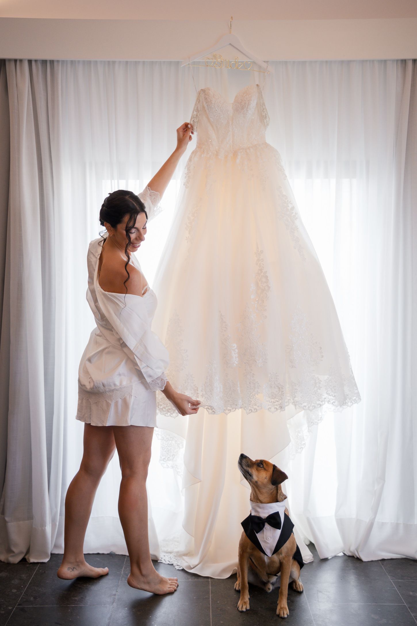 A woman in a robe admires hanging wedding dress while dog in bowtie sits below.