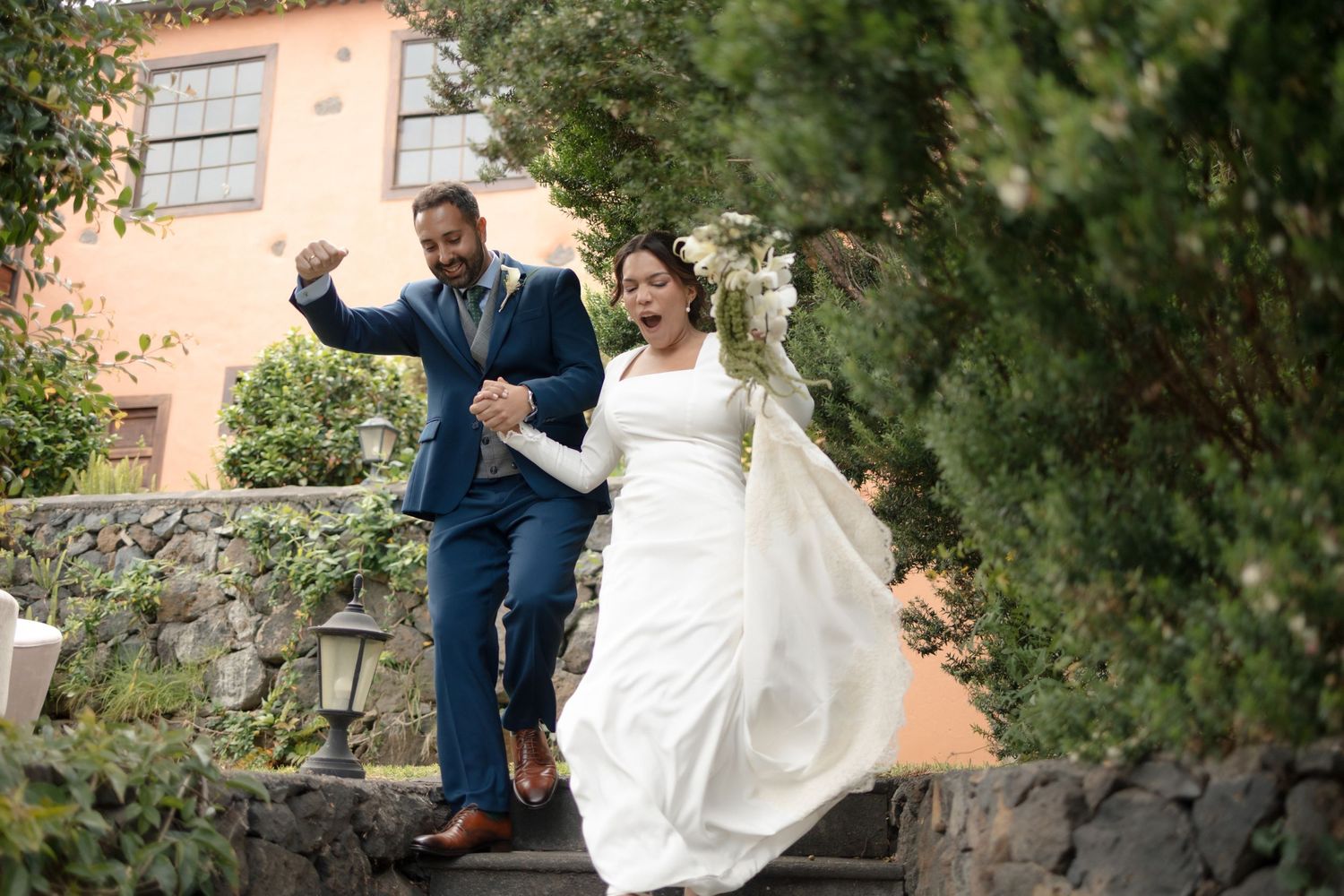 A joyful bride in a white gown and groom in a blue suit hold hands while running up stone steps outdoors.