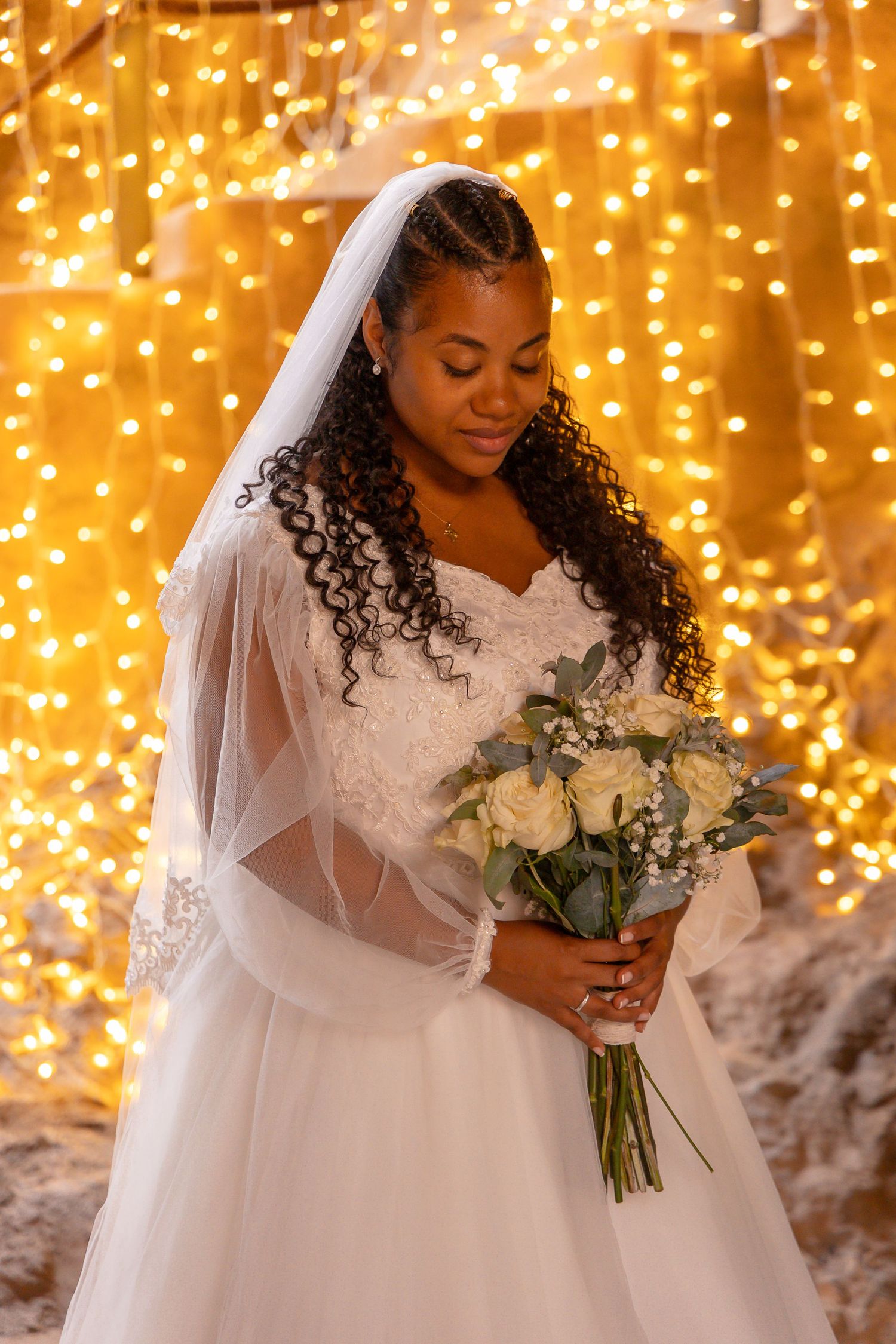 Bride in white dress holding bouquet against backdrop of twinkling golden lights.