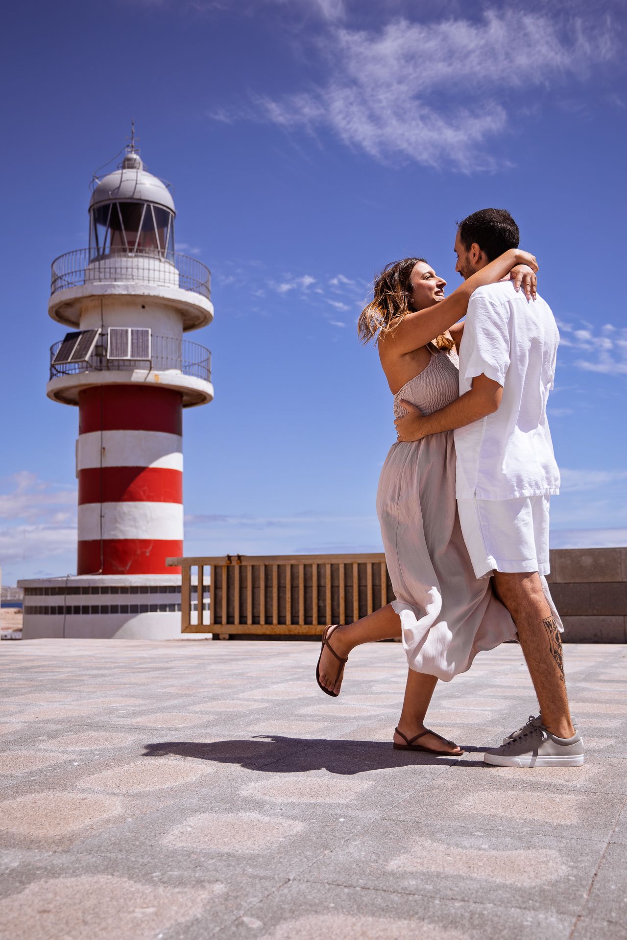 Romantic couple embracing in front of a red and white striped lighthouse against a bright blue sky.