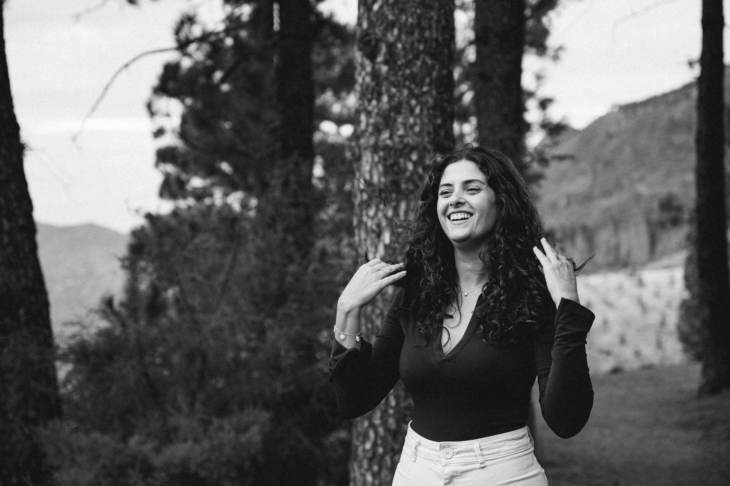 Woman with curly hair laughing joyfully in a black top against a forest backdrop.