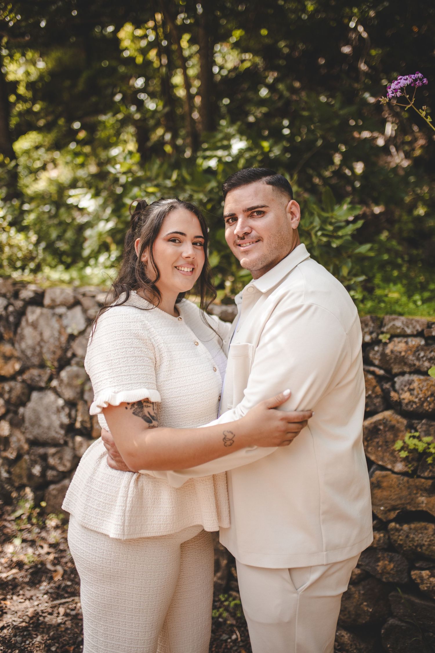 Couple in matching cream outfits embracing outdoors near stone wall with lush green trees, smiling for engagement photo.