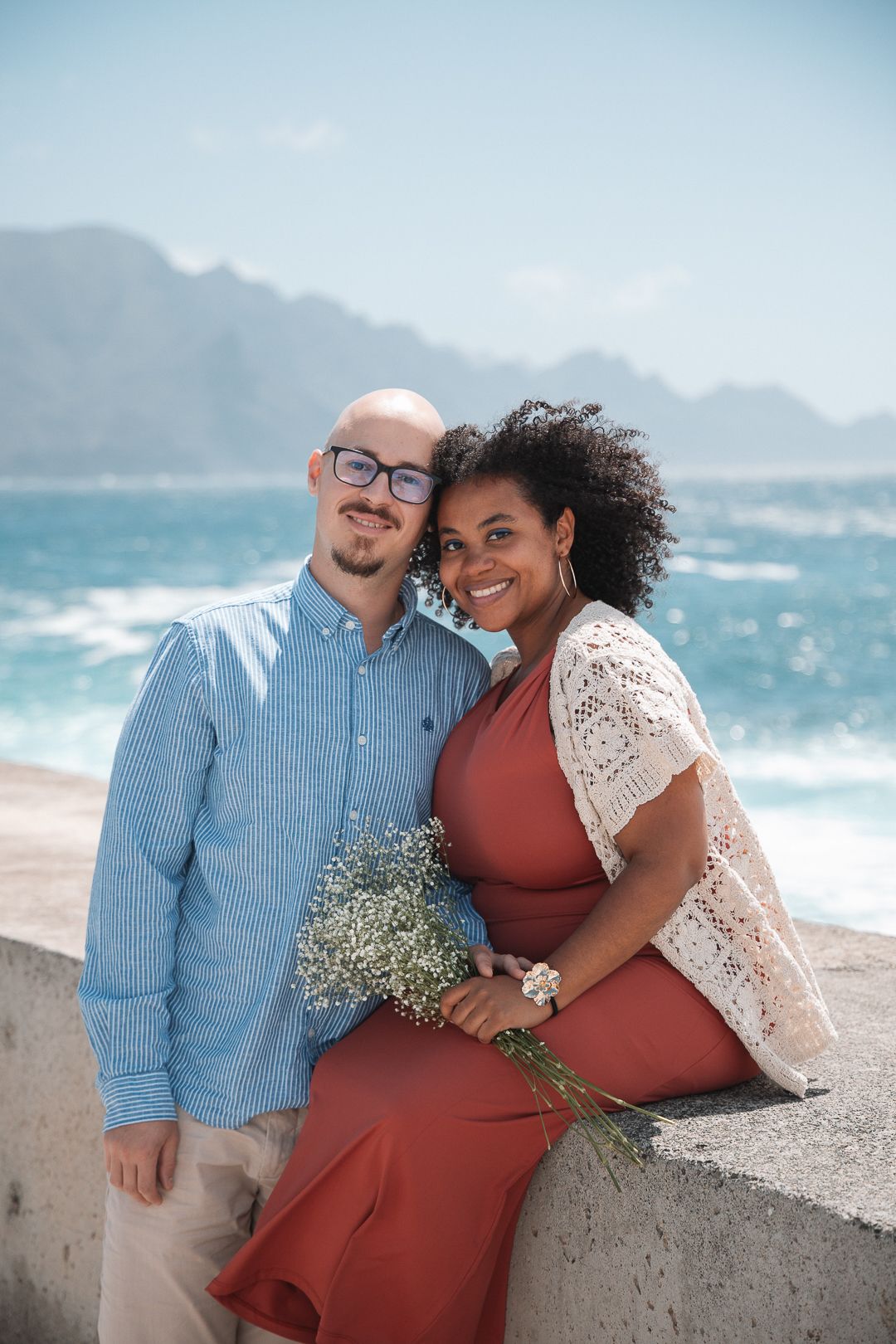 A couple sits together on a concrete wall with crashing ocean waves and mountains in the background.