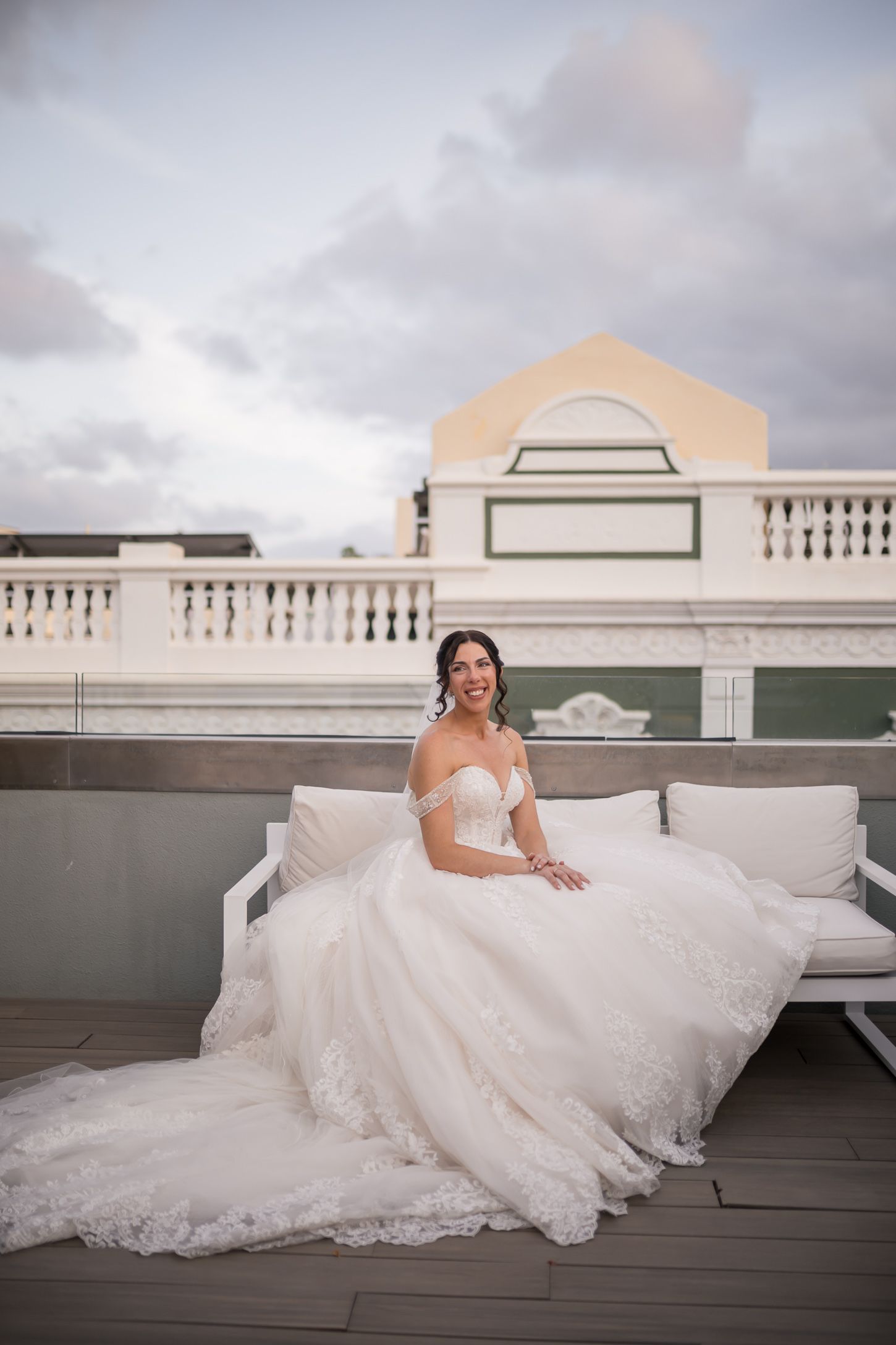 A bride in a flowing lace gown sits elegantly on a rooftop bench at dusk.