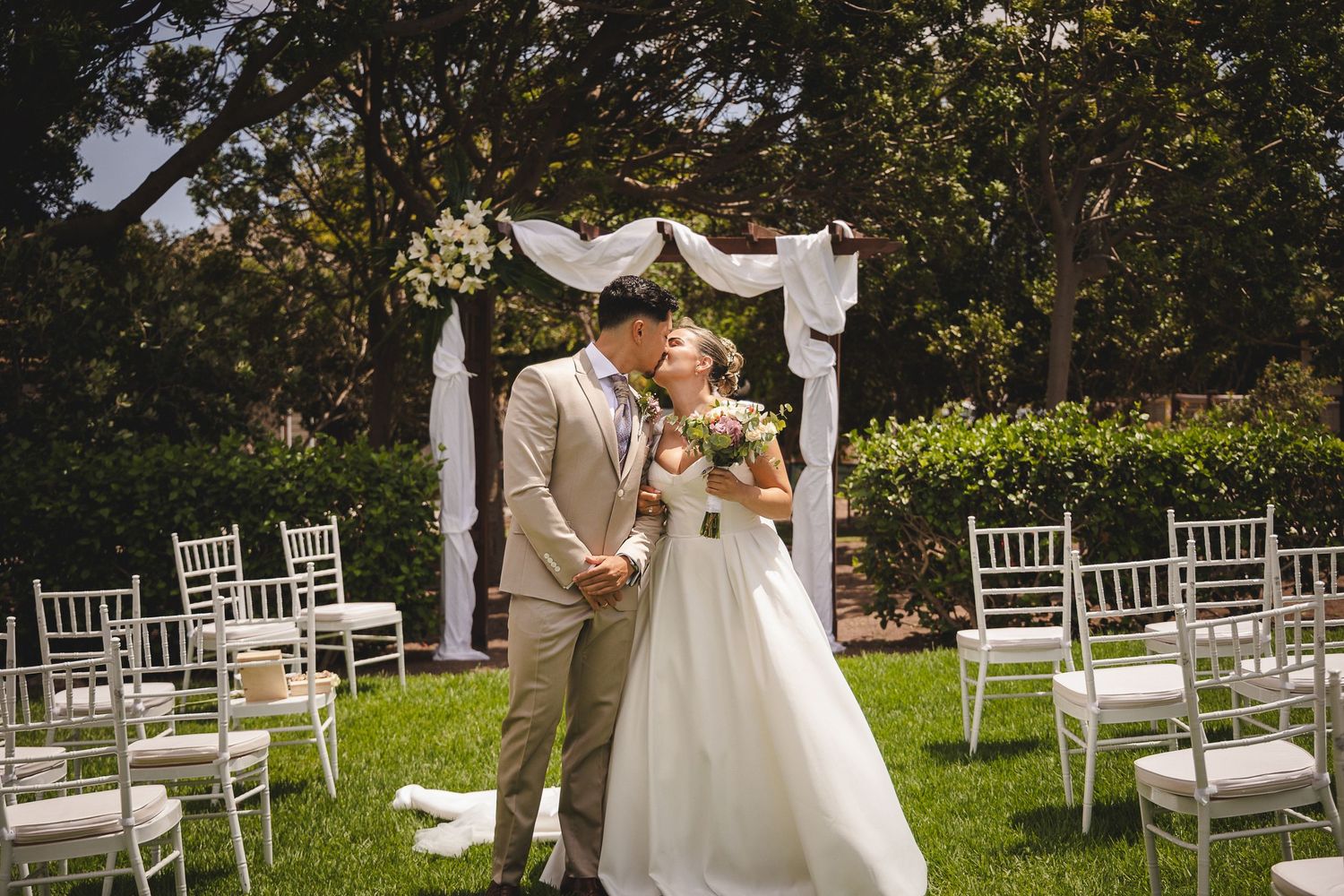 A garden wedding ceremony setup with white chairs and floral arrangements on a sunny day.