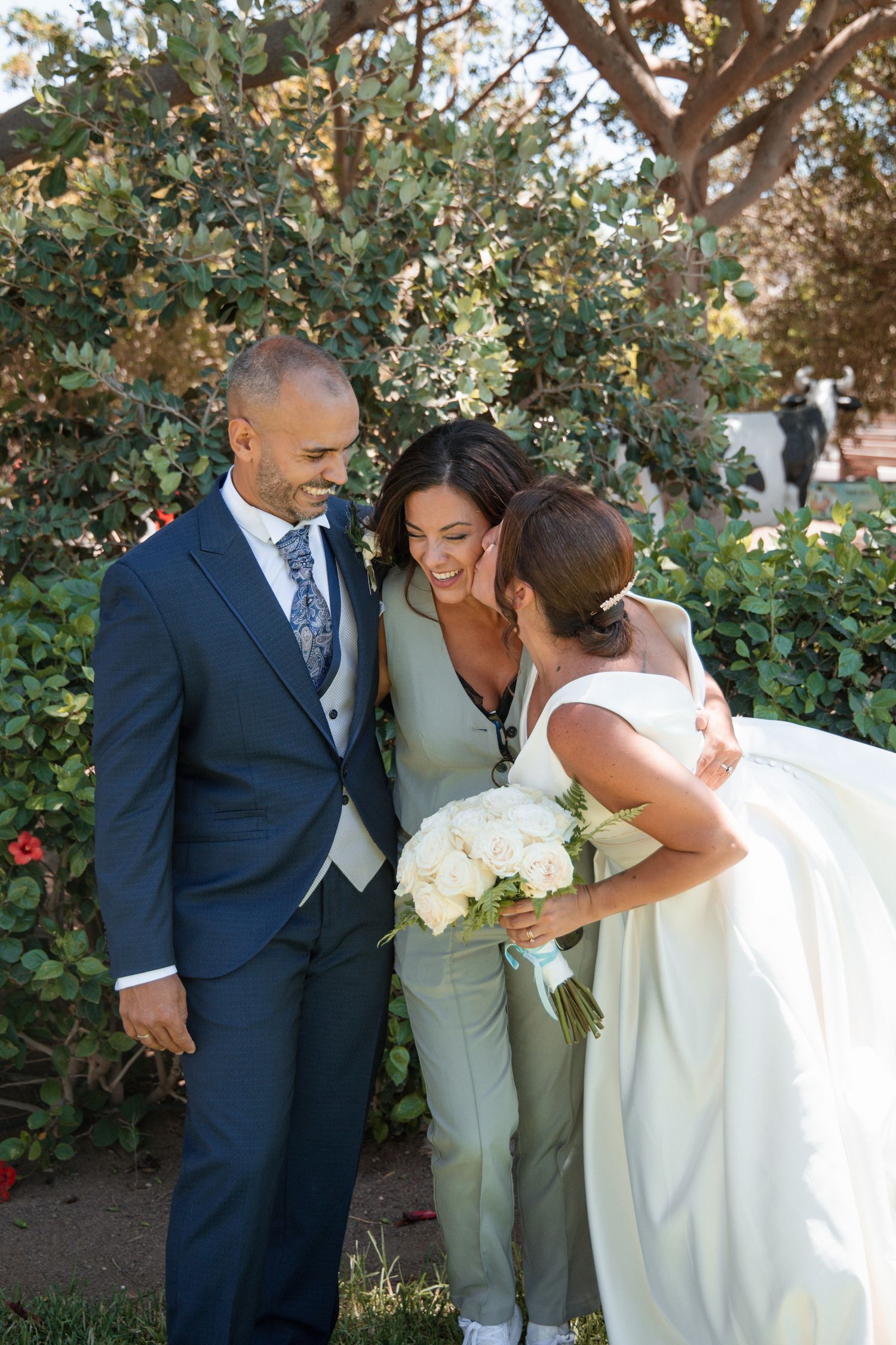A joyful wedding moment shared between three people in formal attire with white bouquet in a garden setting.