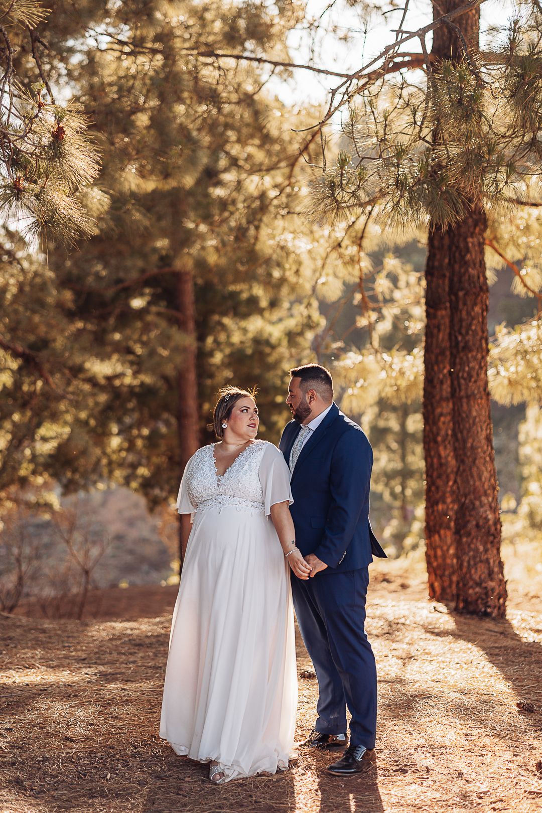 Couple in wedding attire standing among tall pine trees in golden afternoon light.