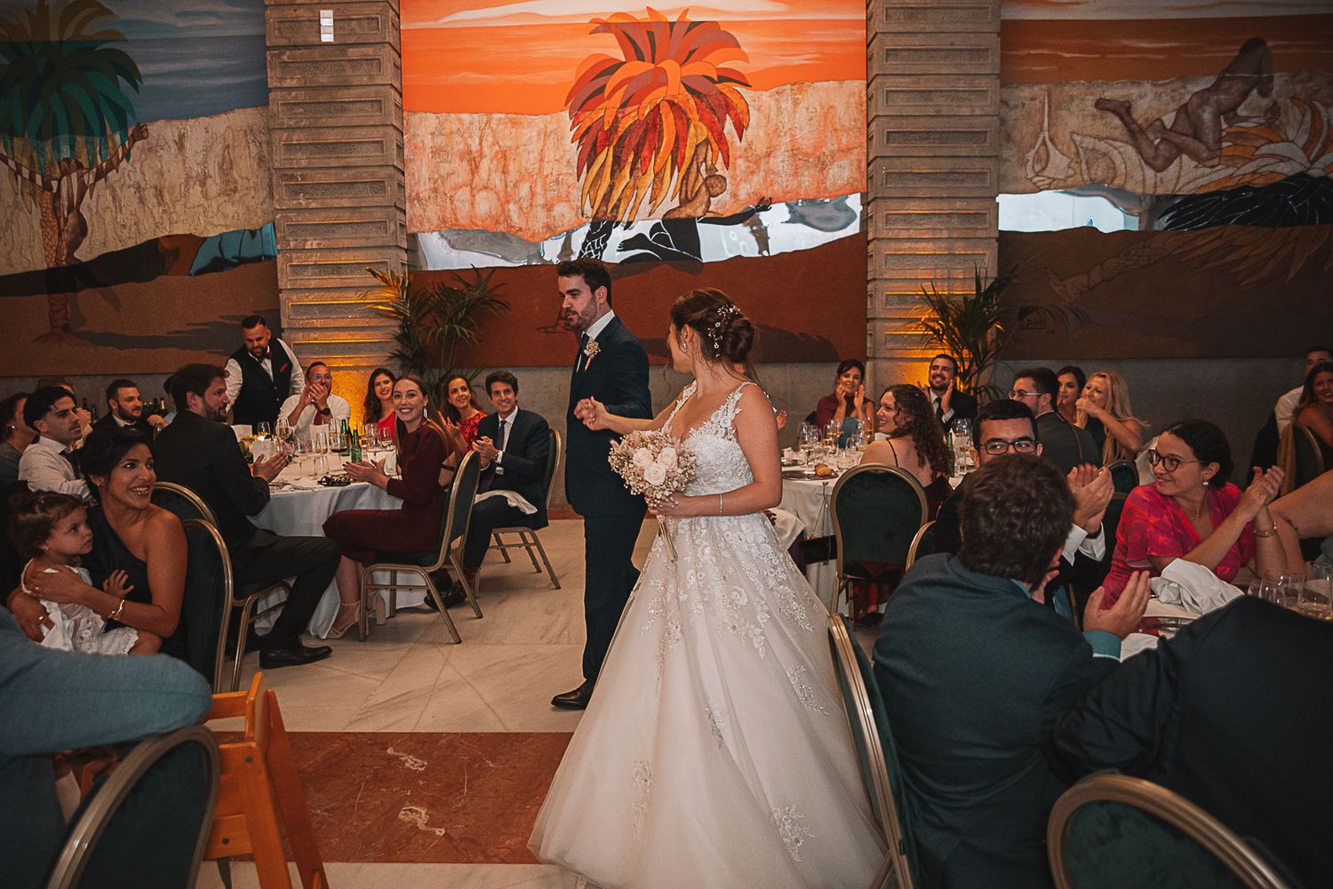 Wedding reception in a warm-lit venue with rustic orange walls and palm decor.