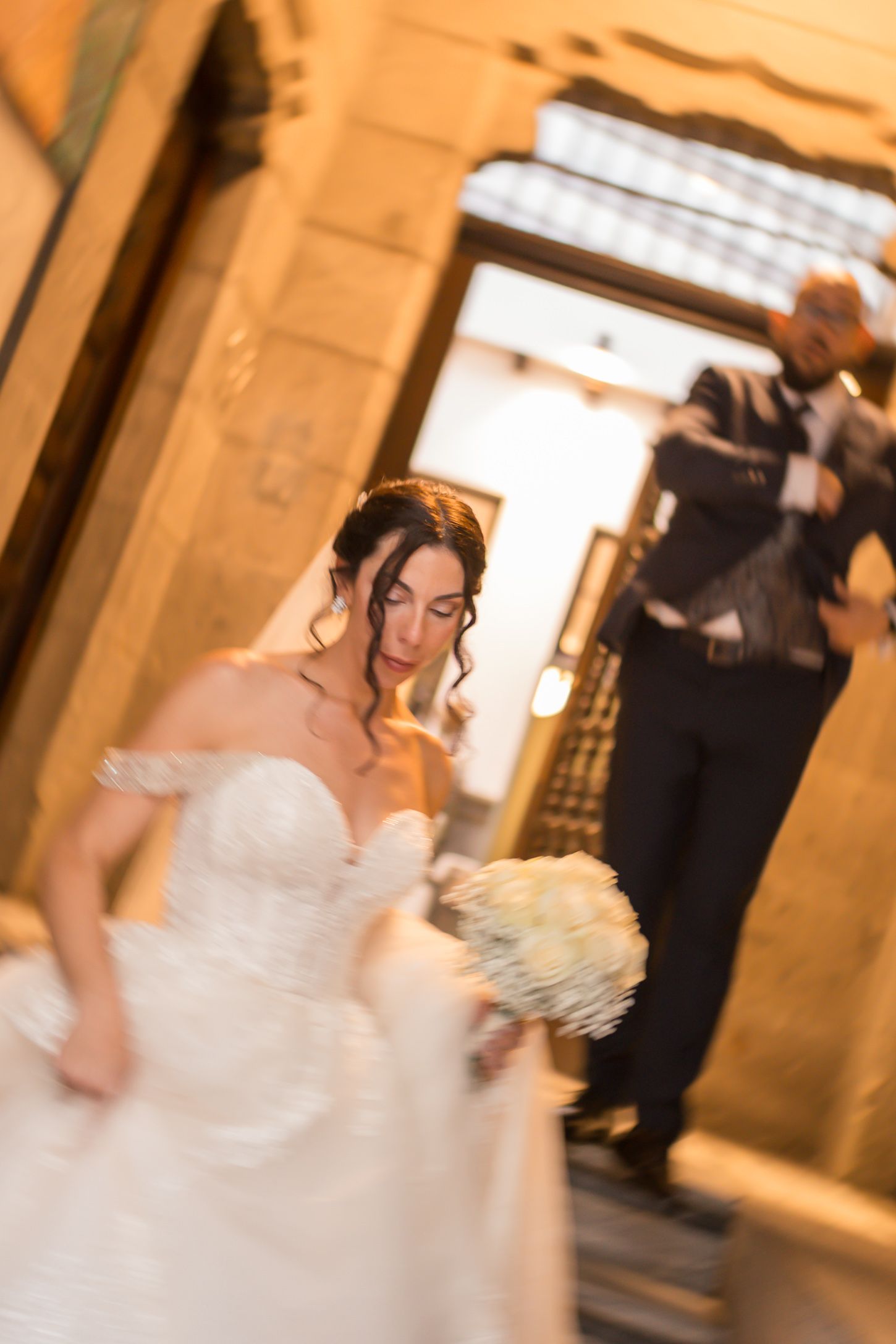 A bride in a white gown holds a bouquet in a warmly lit indoor setting.