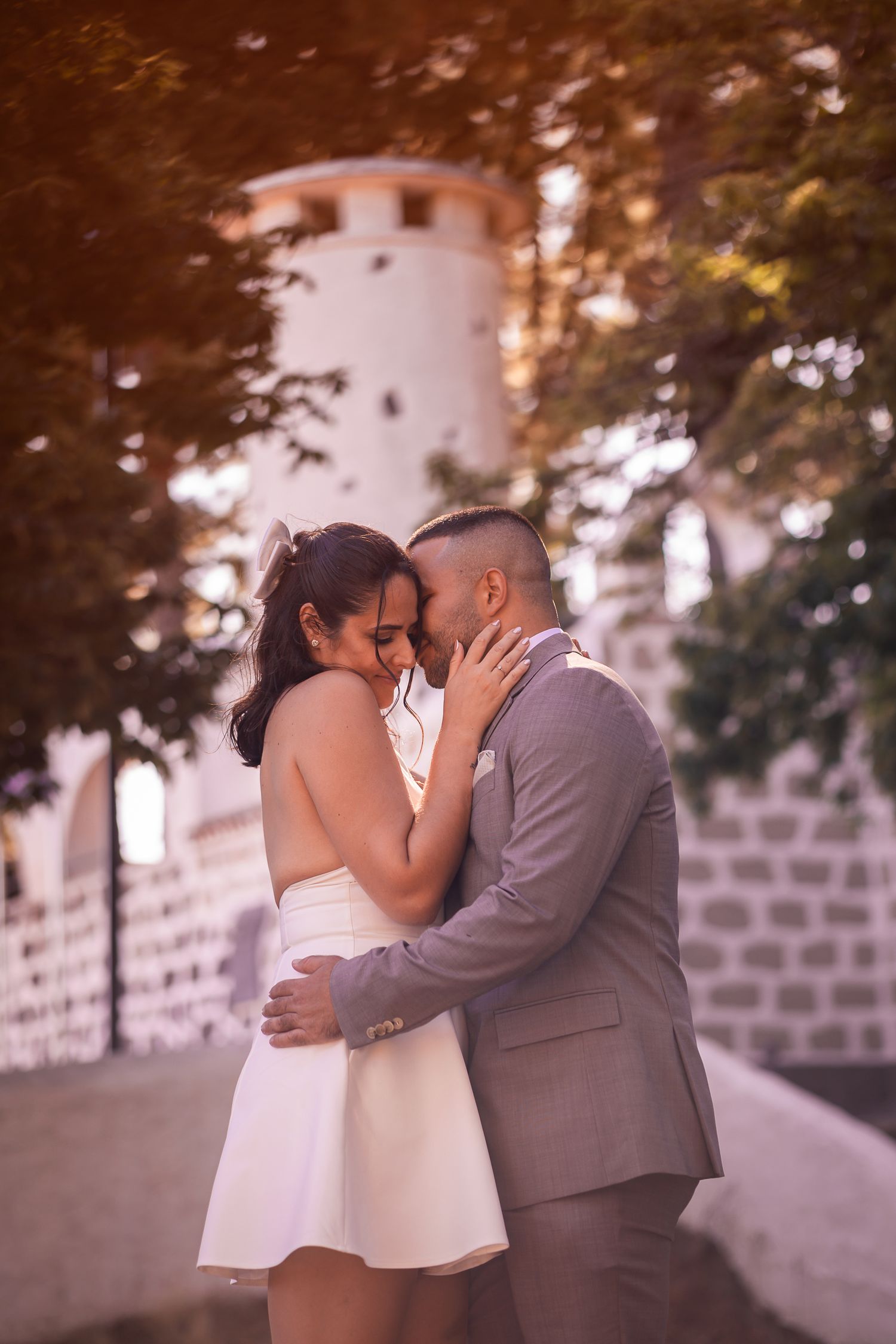A romantic couple in formal attire share an intimate embrace against a stone wall during a sunset photoshoot.