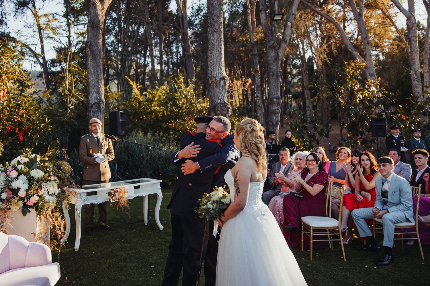 Outdoor wedding reception under trees with guests seated at decorated tables.