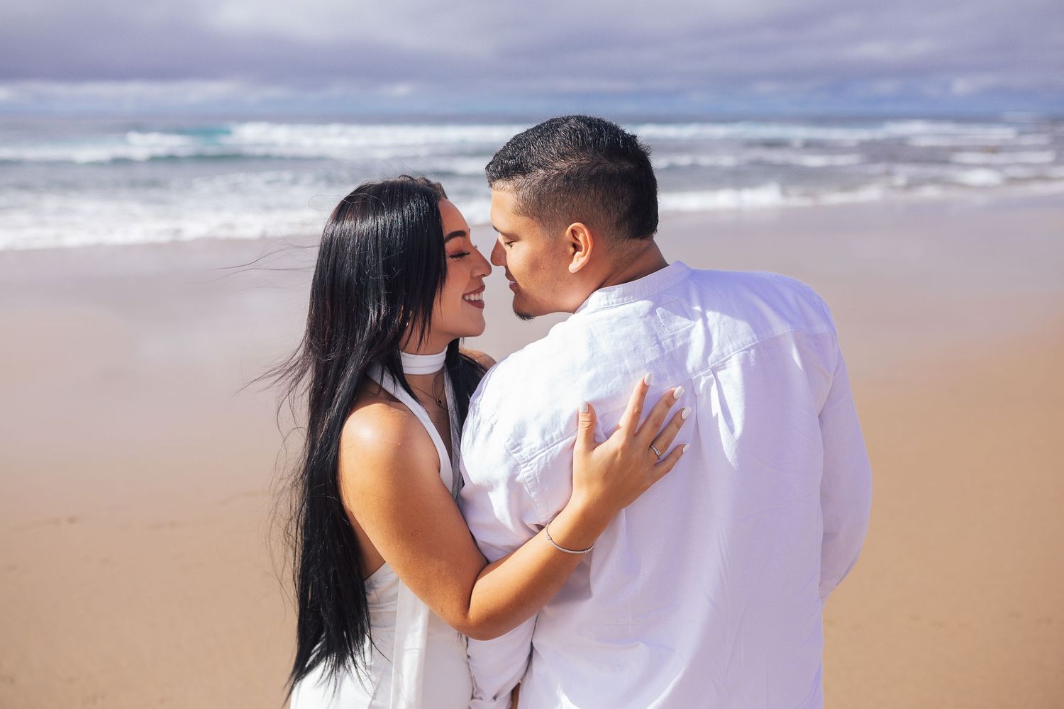Romantic couple in white outfits embracing on a scenic beach during sunset.