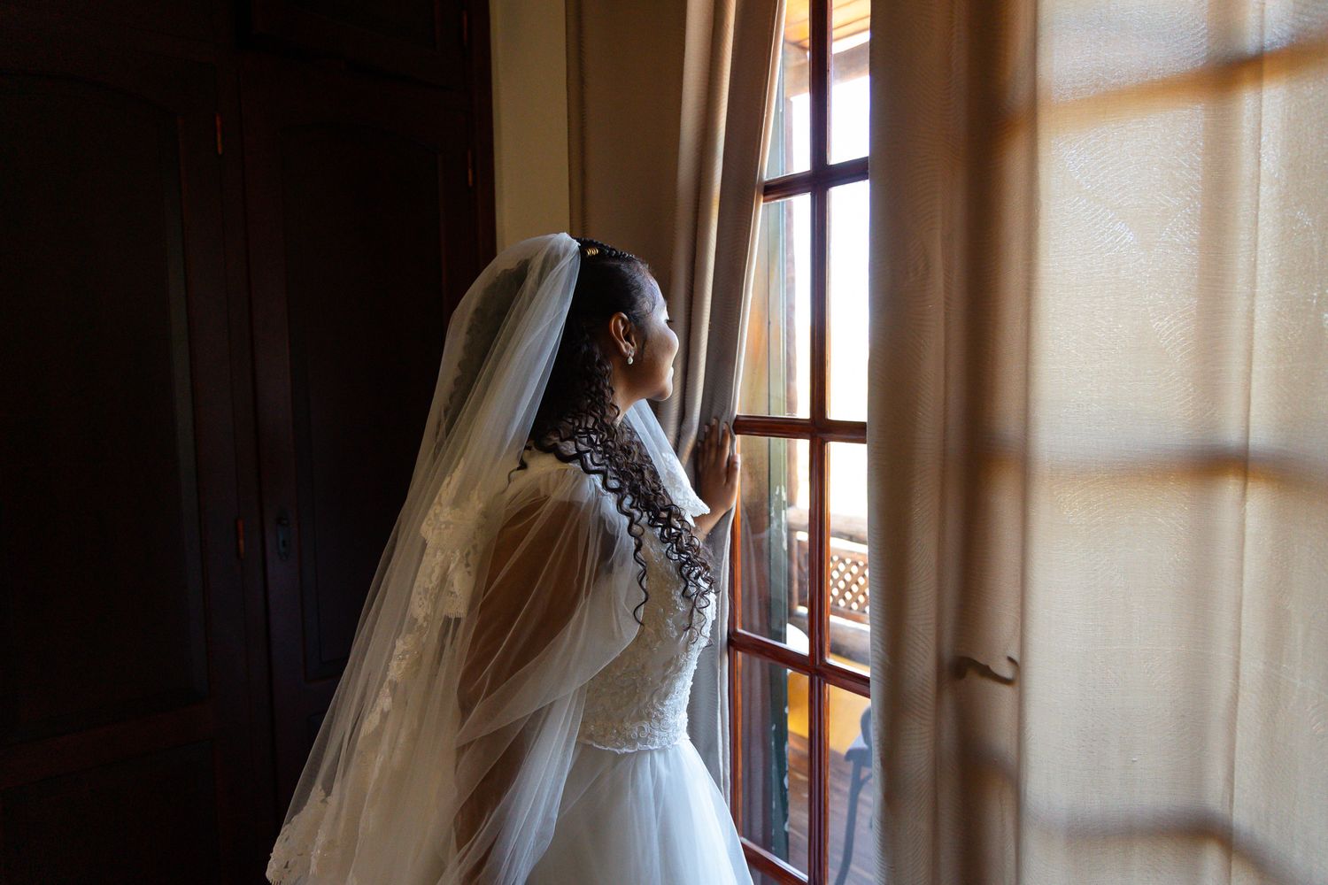 Sunlit bridal preparation scene through window with flowing white dress.