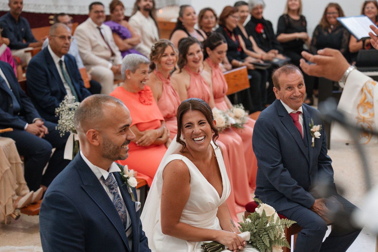 Bride and groom laughing joyfully during wedding ceremony with guests seated in church pews in background.