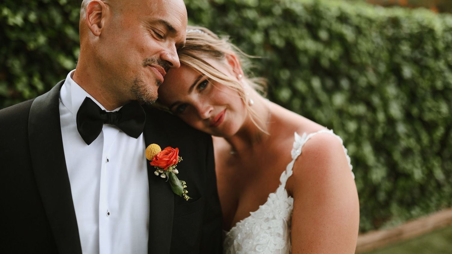 A tender moment between newlyweds at their wedding, with the groom wearing a black tuxedo and red boutonniere.