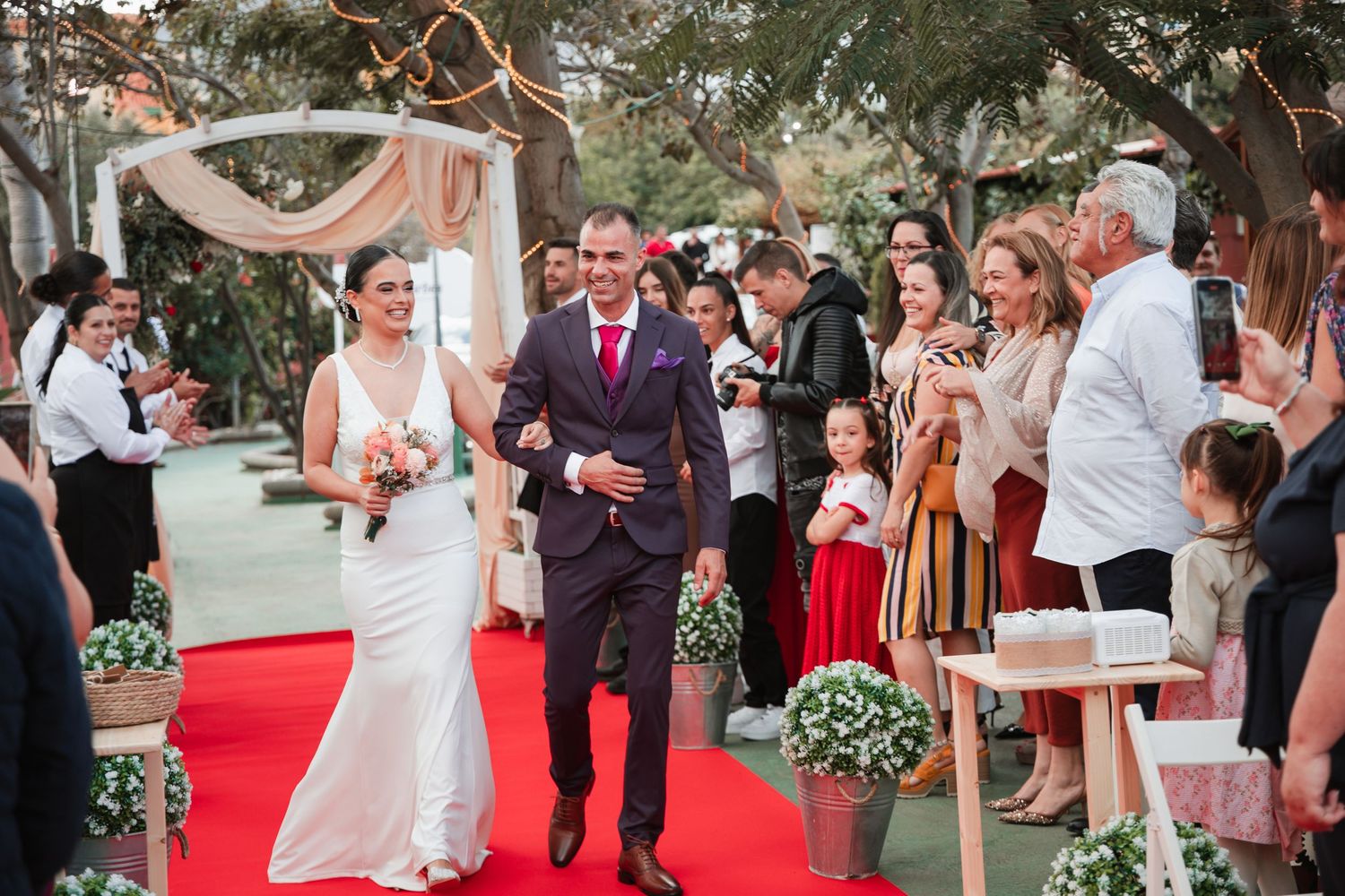 Wedding ceremony guests seated on white chairs along a red carpet aisle outdoors.