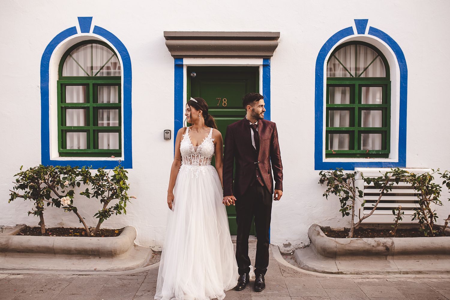 A couple in wedding attire stands in front of a white building with blue-trimmed arched windows and doorways.