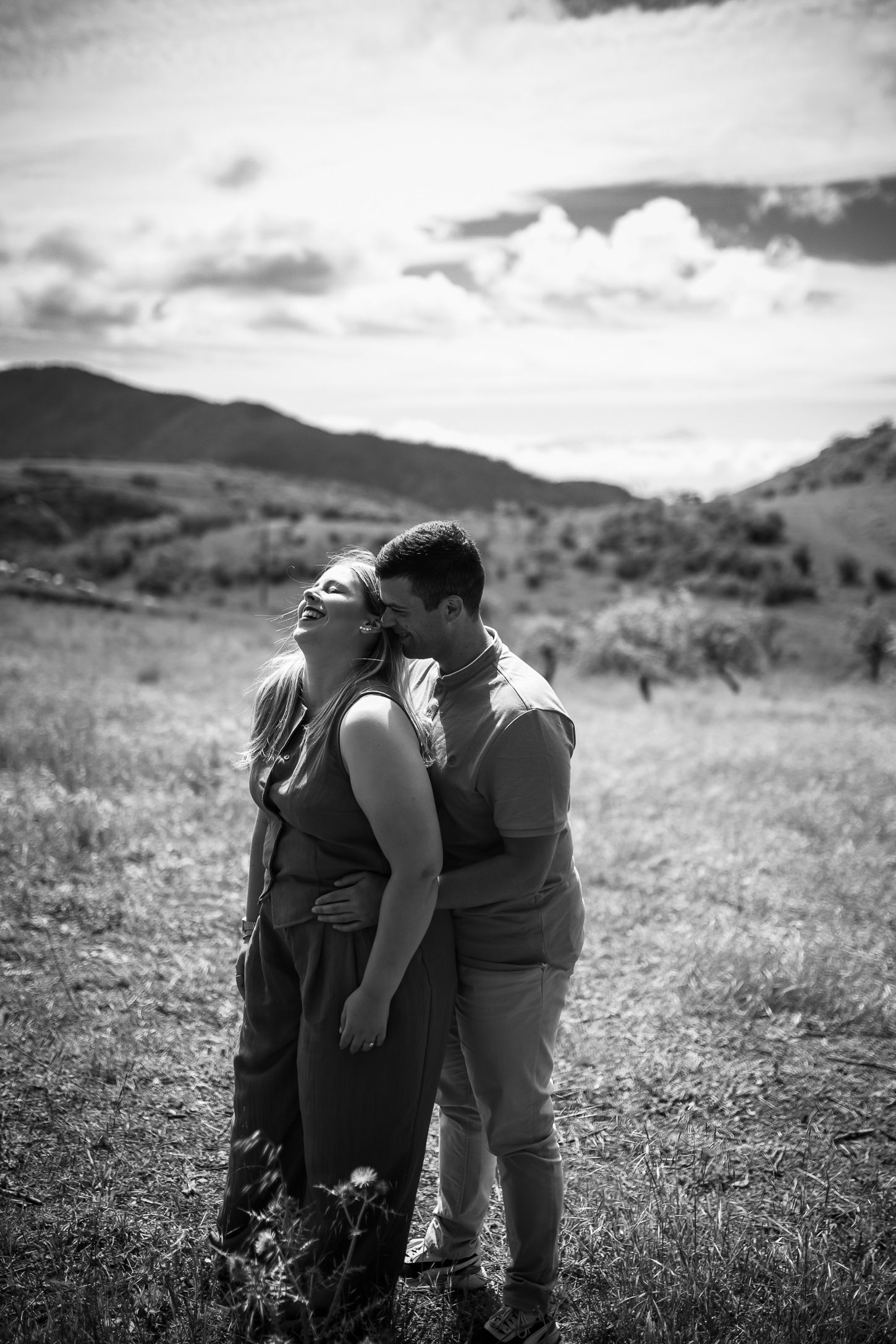 A romantic couple embraces in a grassy field with mountains and dramatic clouds in the background.