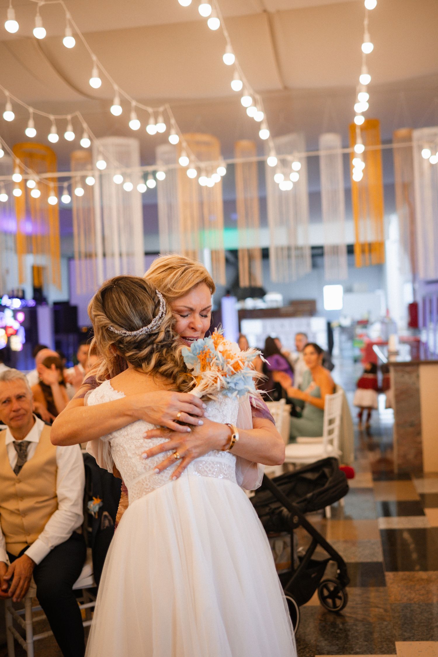 Two people share an emotional embrace on a dance floor under glowing string lights at an indoor wedding reception.