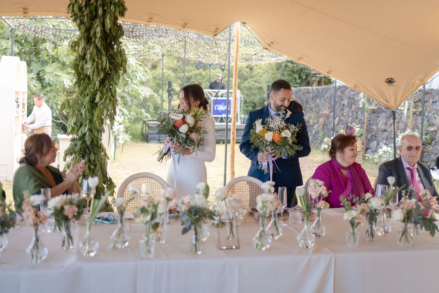 Bride and groom stand at wedding reception head table holding bouquets during speeches under elegant tent.