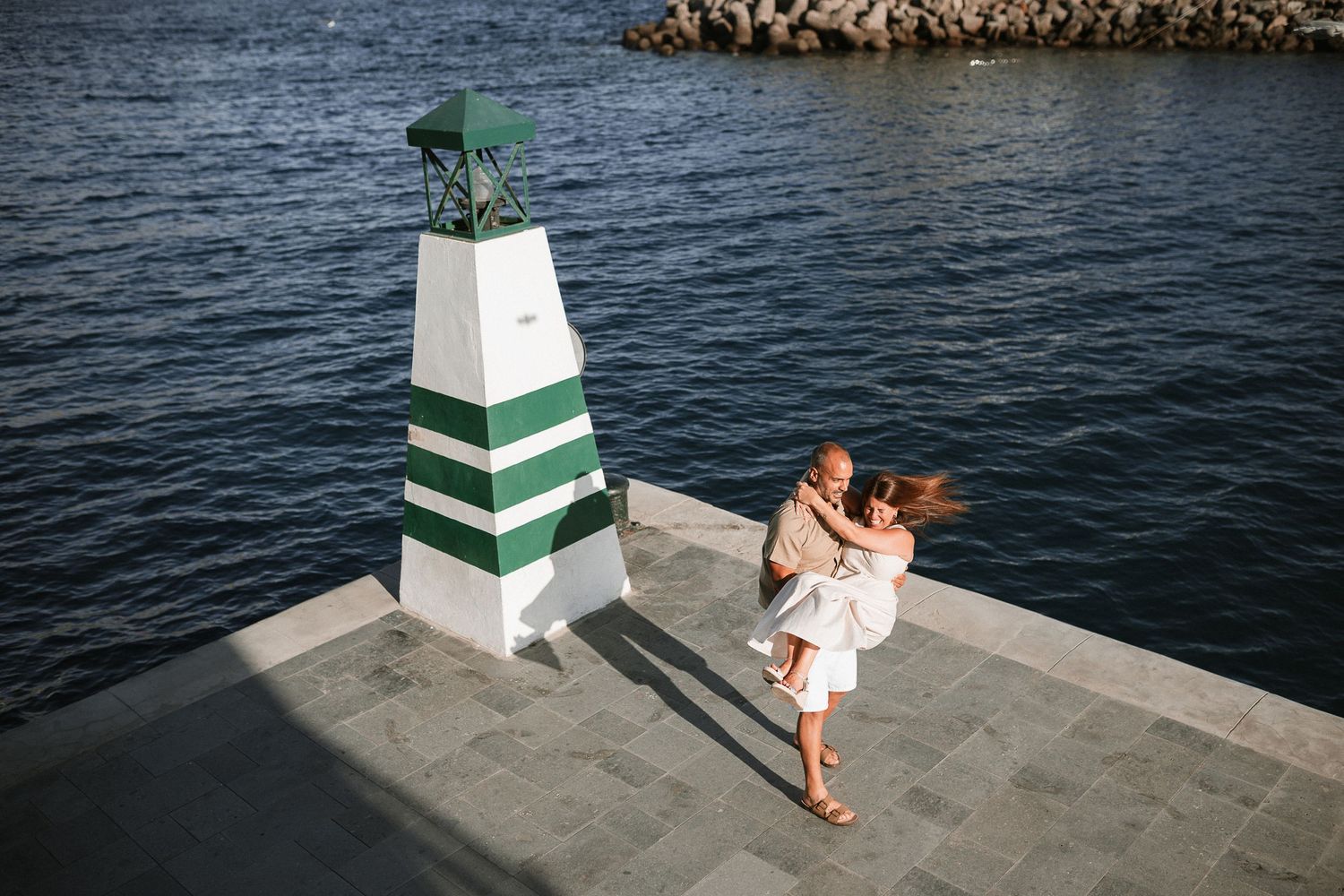 Seaside pier with green and white striped lighthouse against deep blue waters.